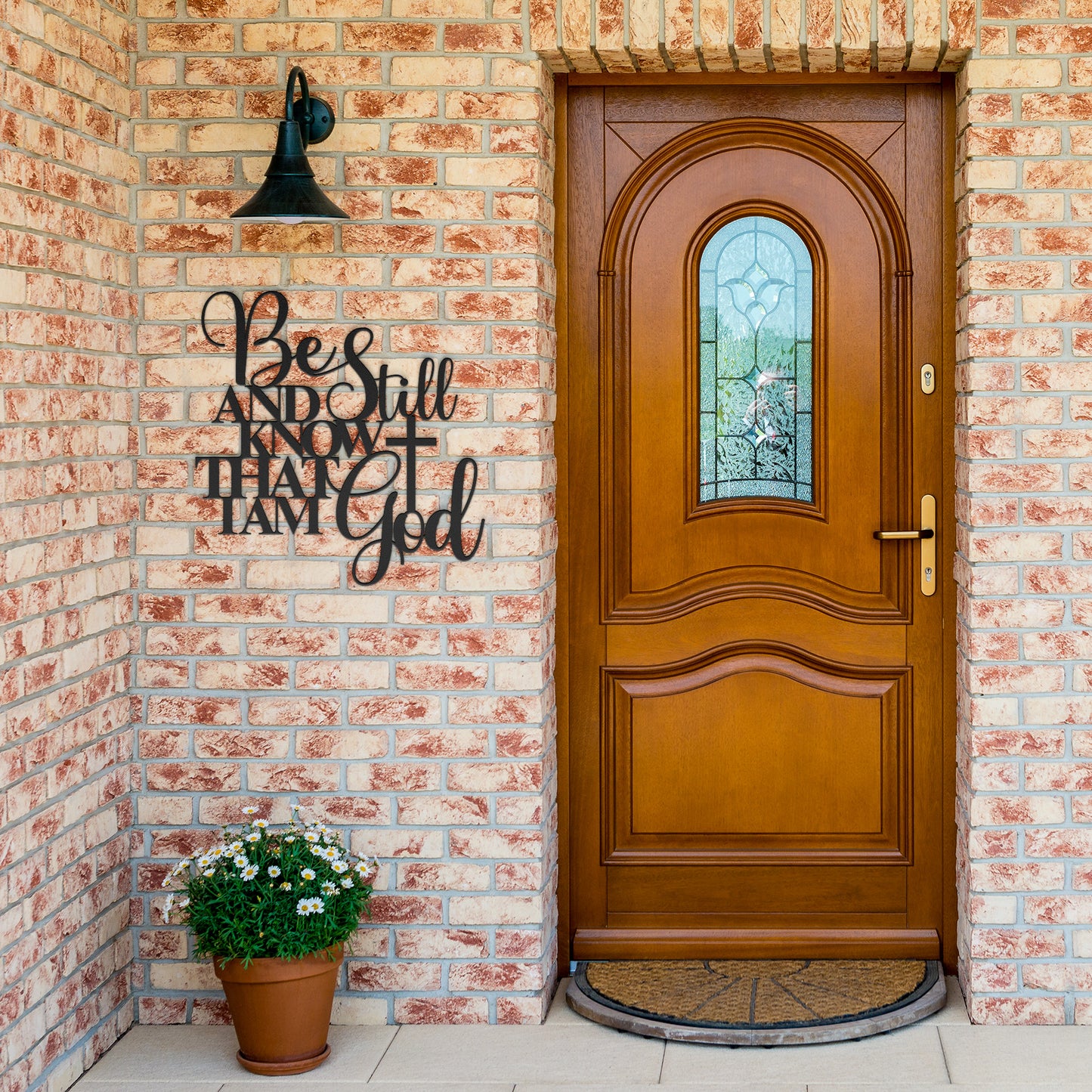A wooden front door with a stained glass panel, brick exterior, a light fixture, and a potted flower beside the entrance. A charming touch is added by the "Be Still And Know Christian Metal Art" sign, giving unique character to the home decor.