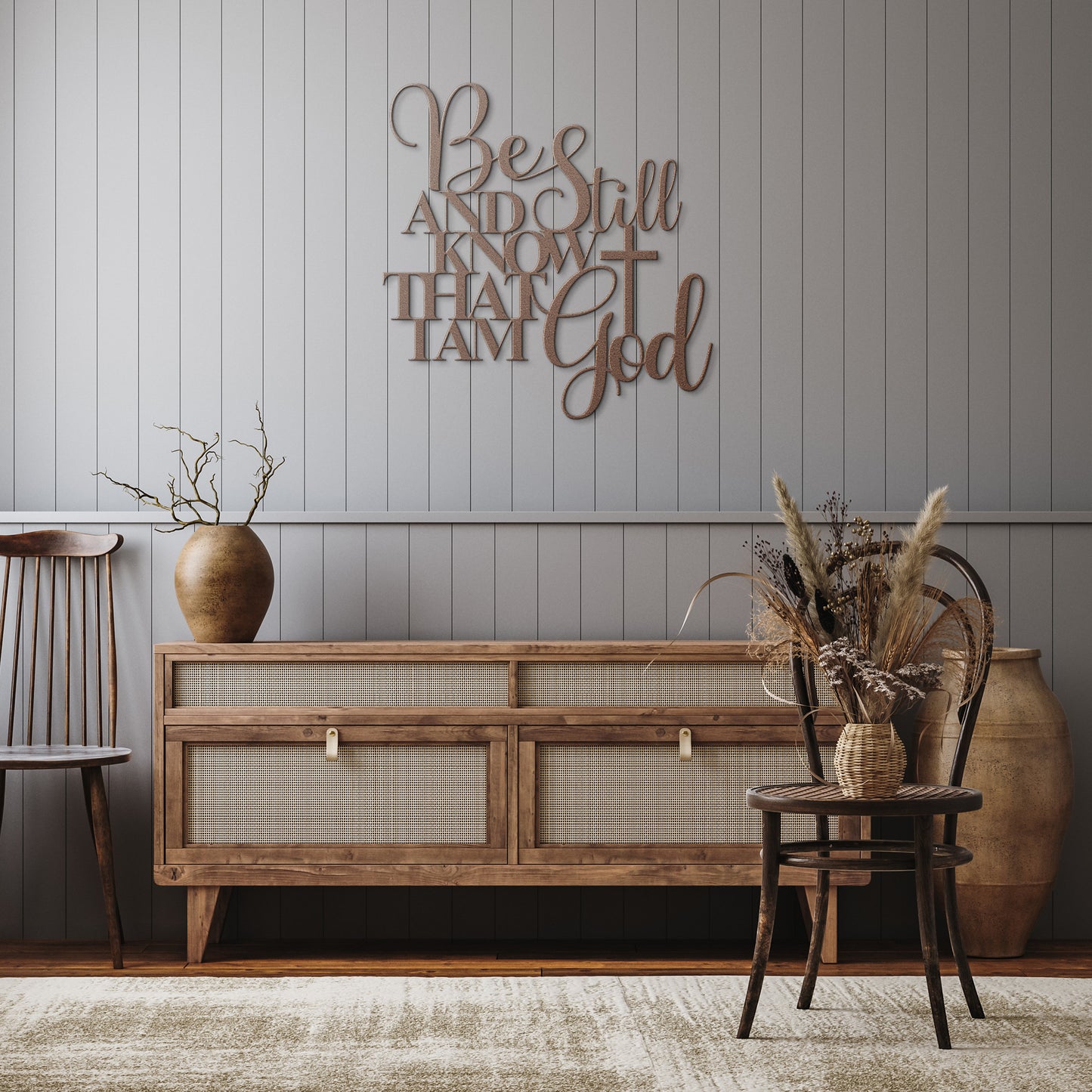 A wooden console table, decorated with vases, stands against a grey paneled wall. Above it, a stylish piece of art named "Be Still And Know Christian Metal Art" reads, "Be still and know that I am God." Two chairs and a rug complete the home decor.