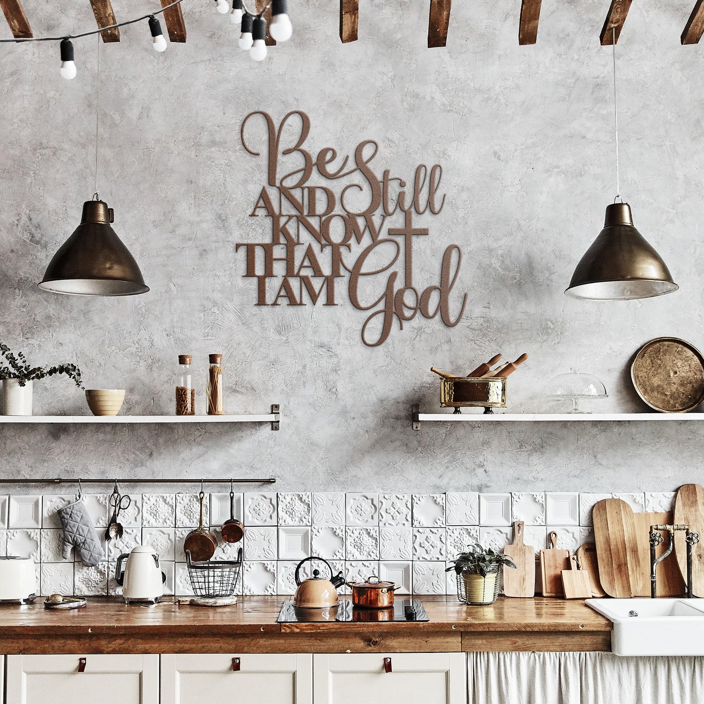 A rustic kitchen with white cabinets, wooden countertops, and shelves features a metal sign called "Be Still And Know Christian Metal Art," crafted from 18 gauge steel. Two pendant lights hang from the ceiling, illuminating the wall decor that displays the text "Be Still and Know That I am God.