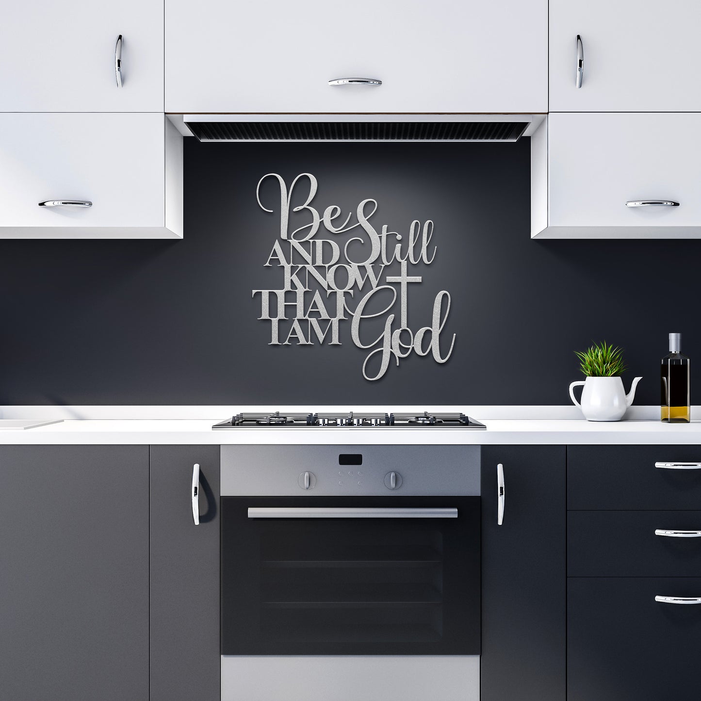 Modern kitchen with white cabinets, black countertops, and an oven. Above the stove hangs the "Be Still And Know Christian Metal Art," which features a cross and reads, "Be Still and Know that I am God." A small plant and oil bottle on the counter add a touch of natural home decor.