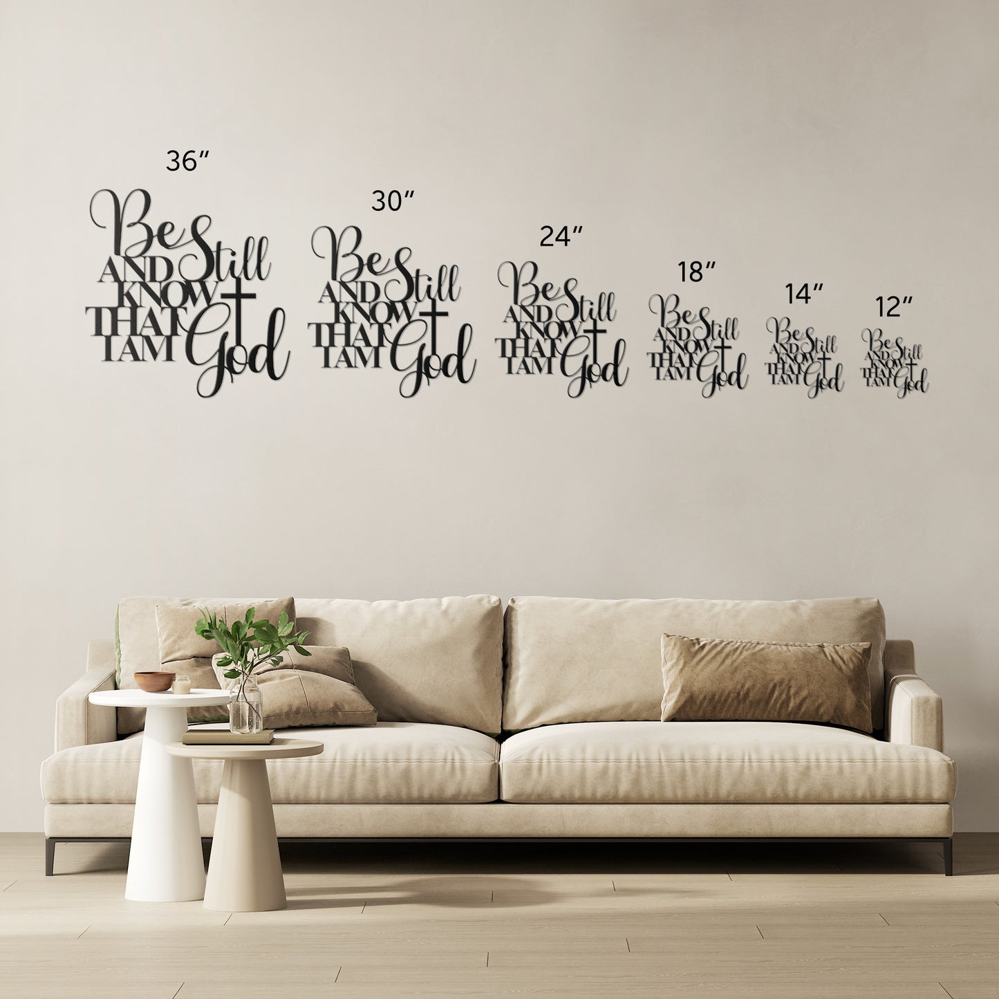 A beige couch sits against a white wall adorned with the "Be Still And Know Christian Metal Art," featuring multiple decals of varying sizes that read, "Be still and know that I am God." Beside the couch, a small round table with a plant and lamp completes the home decor, adding charm and coziness to the space.