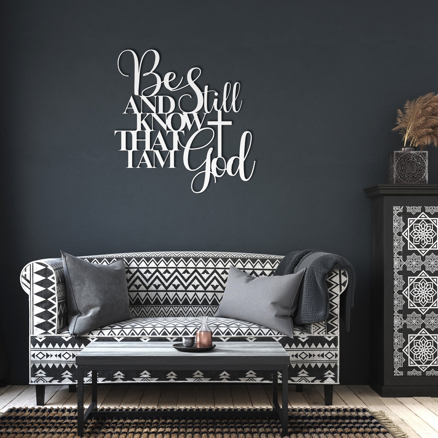 A black and white patterned sofa with cushions is in front of a dark grey wall adorned with the "Be Still And Know Christian Metal Art," an 18 gauge steel metal sign featuring a decorative religious quote. A small black coffee table and a patterned cabinet filled with decorative items complete the home decor scene.