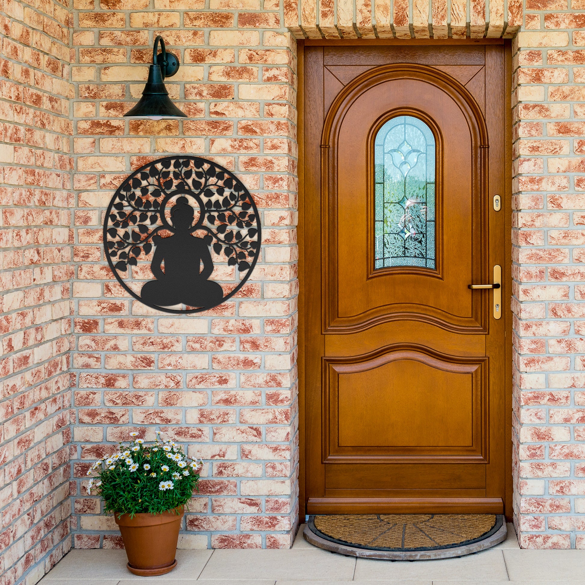 A wooden door with an arched window and brass handle is set in a brick wall. A round, powder-coated metal sign featuring the Buddhist Mandala Metal Art depicting a meditating figure under a tree and a potted plant are next to the door, adding charm to the home decor.