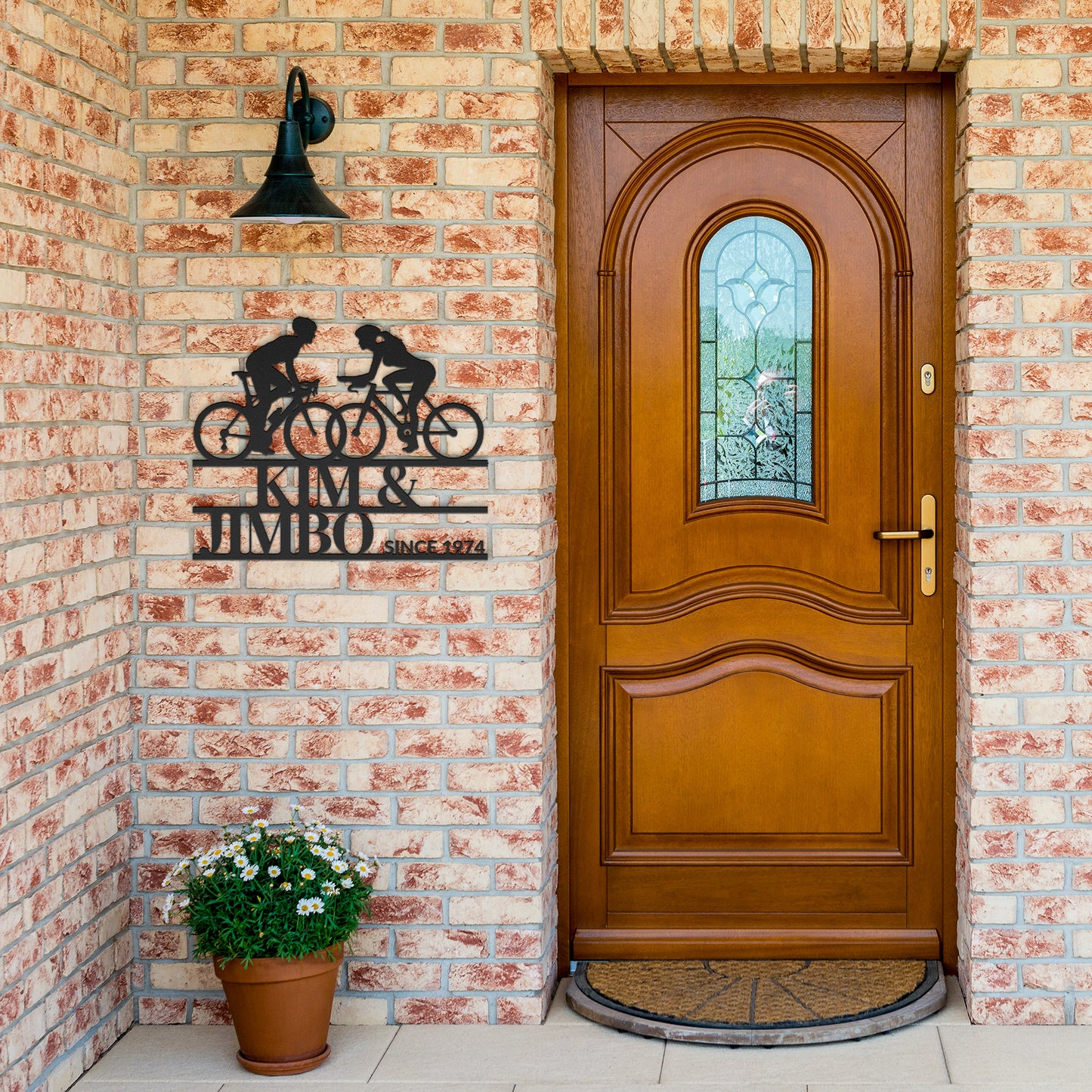 A wooden door with an arched window is set in a brick wall. On the wall hangs a custom metal wall art piece called "Cyclist Couple Metal Wall Art Sign," alongside a black lamp. A potted plant sits on the ground near the door.