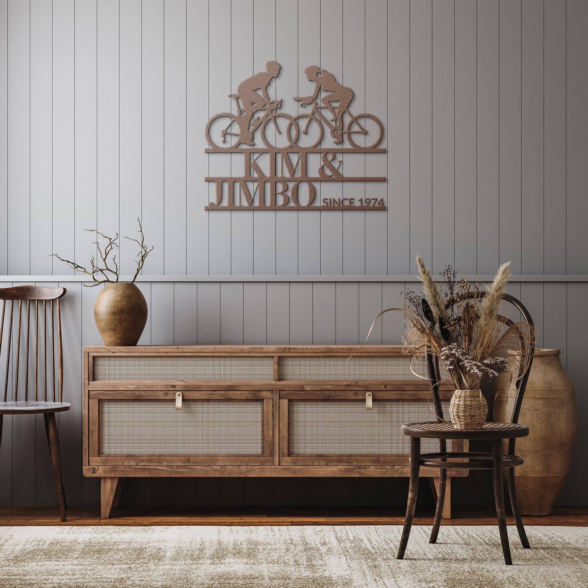 A rustic room features a wooden sideboard, two chairs adorned with vases and dry plants, and the striking Custom Cyclist Couple Metal Wall Art Sign. This personalized decor piece is engraved with "KIM & JIMBO SINCE 1974," adding charm to any space as a unique couple's gift or cycling-themed artwork.