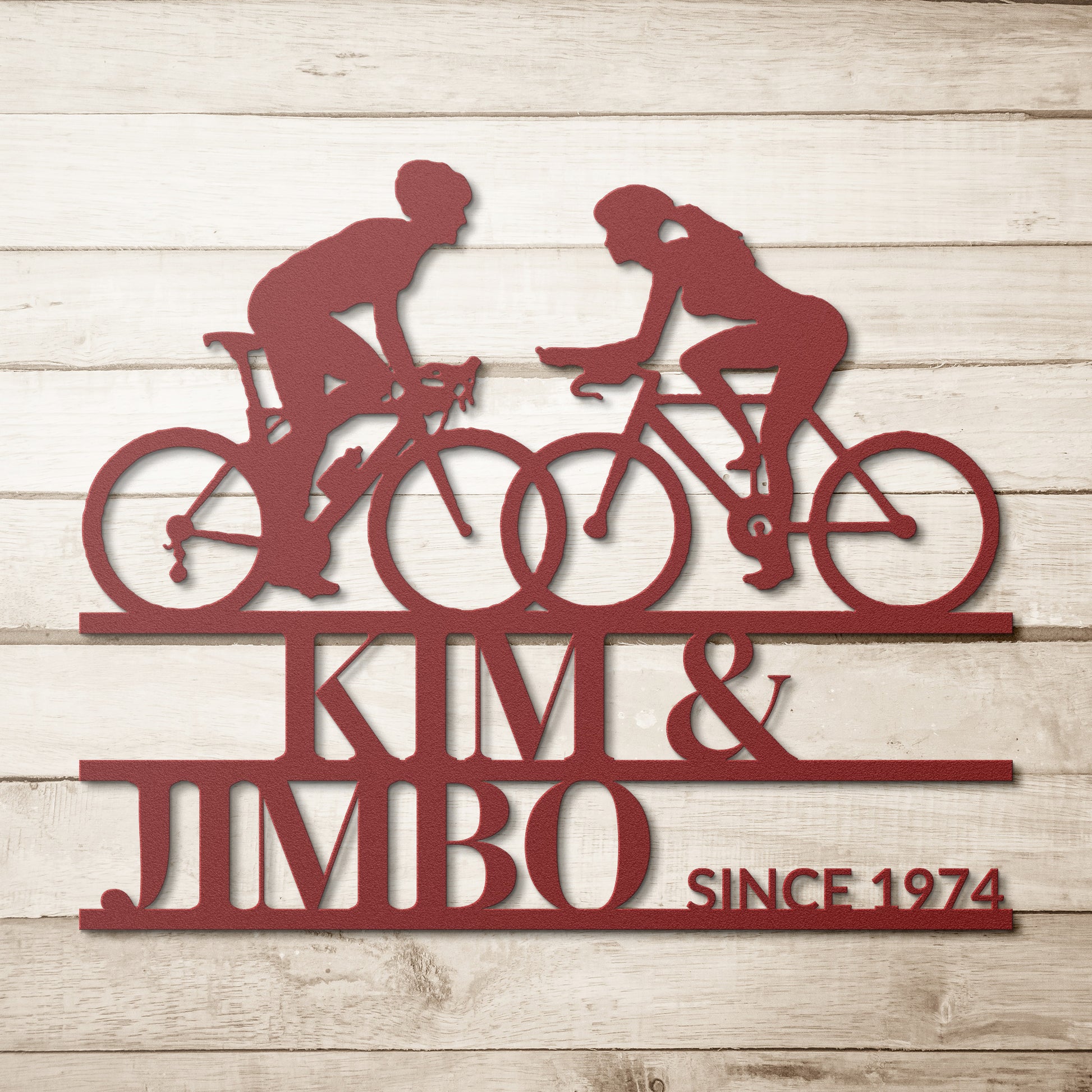 Custom Cyclist Couple Metal Wall Art Sign featuring a metal silhouette of two cyclists with the text "Kim & Jimbo Since 1974" on a wooden backdrop, making it an ideal personalized home decor gift.