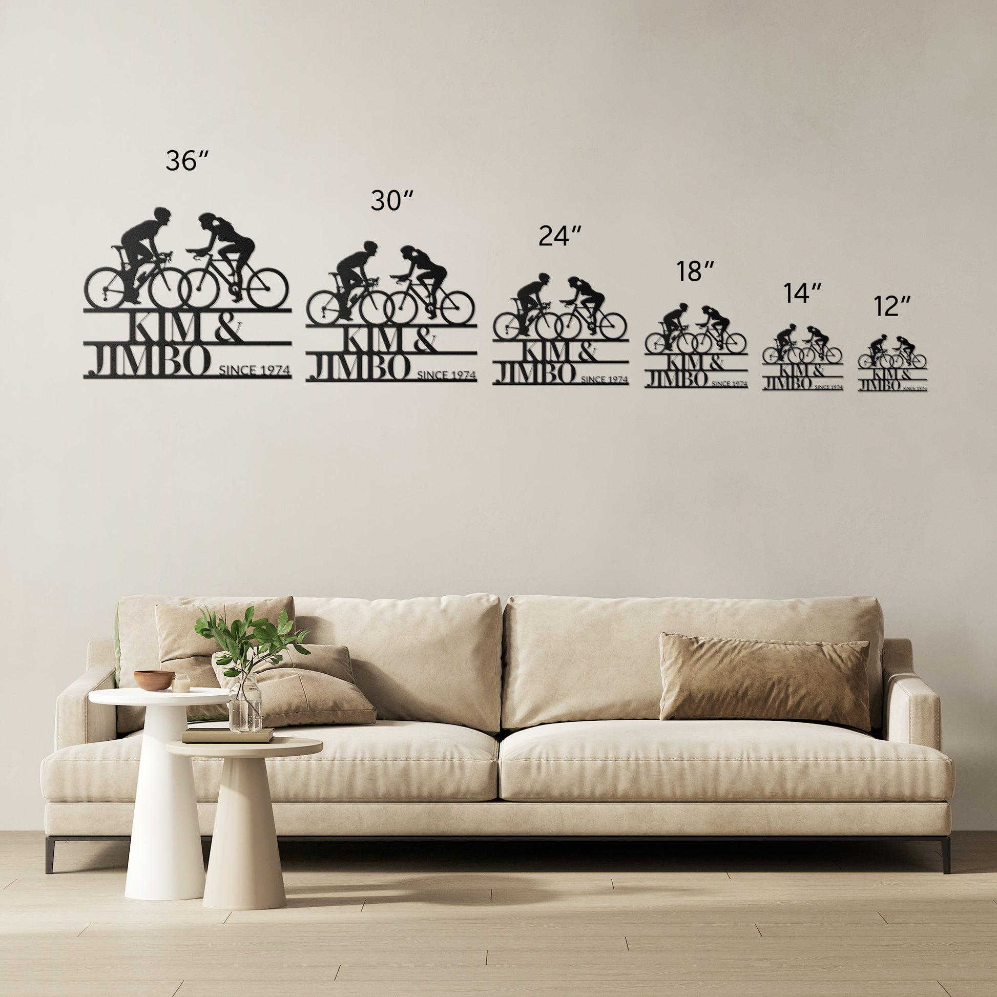 The Custom Cyclist Couple Metal Wall Art Sign, designed with silhouettes of cyclists in varying sizes, is showcased above a beige sofa adorned with cushions. Beside the sofa, a round white table featuring decorative items enhances the ambiance, making it an ideal home decor gift.