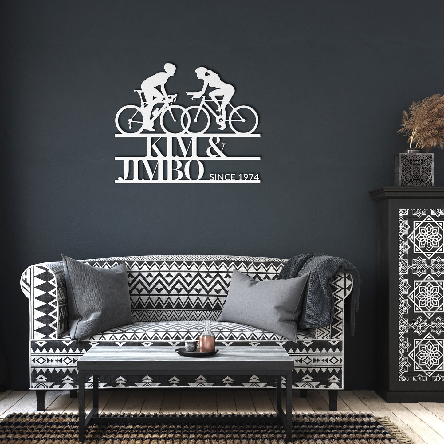 The black and white living room showcases a patterned sofa, cushions, and the striking Custom Cyclist Couple Metal Wall Art Sign: "Kim & Jimbo Since 1974," making it an ideal decor piece or gift for any home.