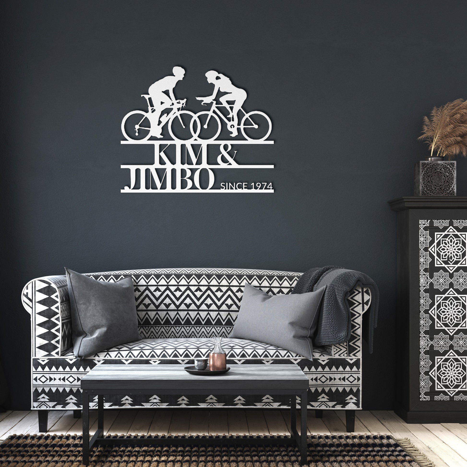 The black and white living room showcases a patterned sofa, cushions, and the striking Custom Cyclist Couple Metal Wall Art Sign: "Kim & Jimbo Since 1974," making it an ideal decor piece or gift for any home.
