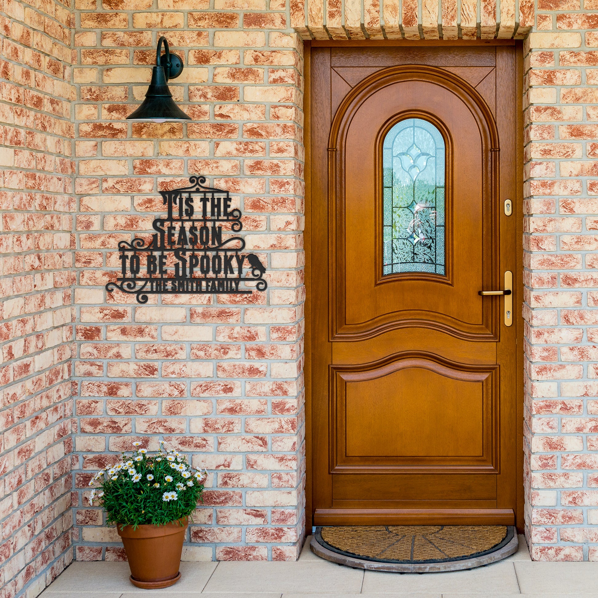 A brick wall with a wooden arched door and a Custom Halloween Metal Sign reading "Tis the season to be spooky." A potted plant with white flowers is placed on the left side of the door.
