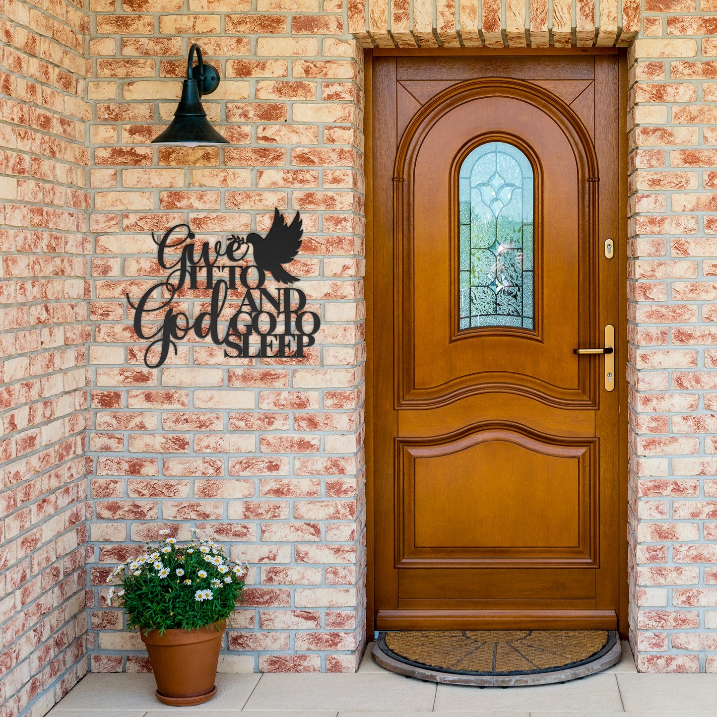 A brick exterior wall with a wooden door featuring a stained glass window. A potted plant is on the left, and a high-quality steel wall sign from the collection "Give It To God And Go To Sleep Christian Metal Sign" reads those very words, adding an inspirational touch of Christian bedroom wall art and decor.