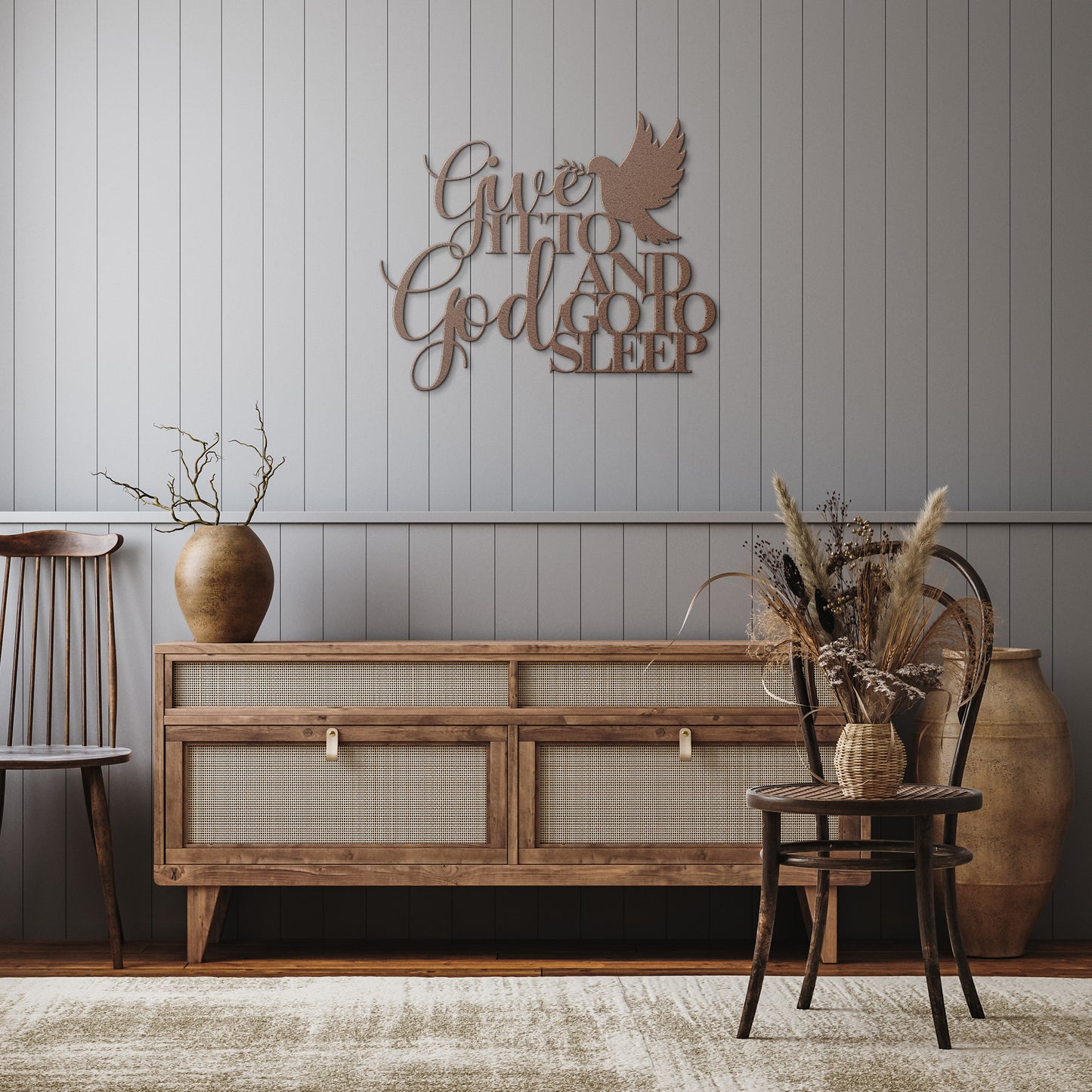 A wooden sideboard with a vase of dried flowers sits against a gray, paneled wall adorned with the "Give It To God And Go To Sleep" Christian Metal Sign—an inspirational quote that enhances your home decor. Flanked by two wooden chairs, this piece adds a meaningful touch to the room's ambiance.