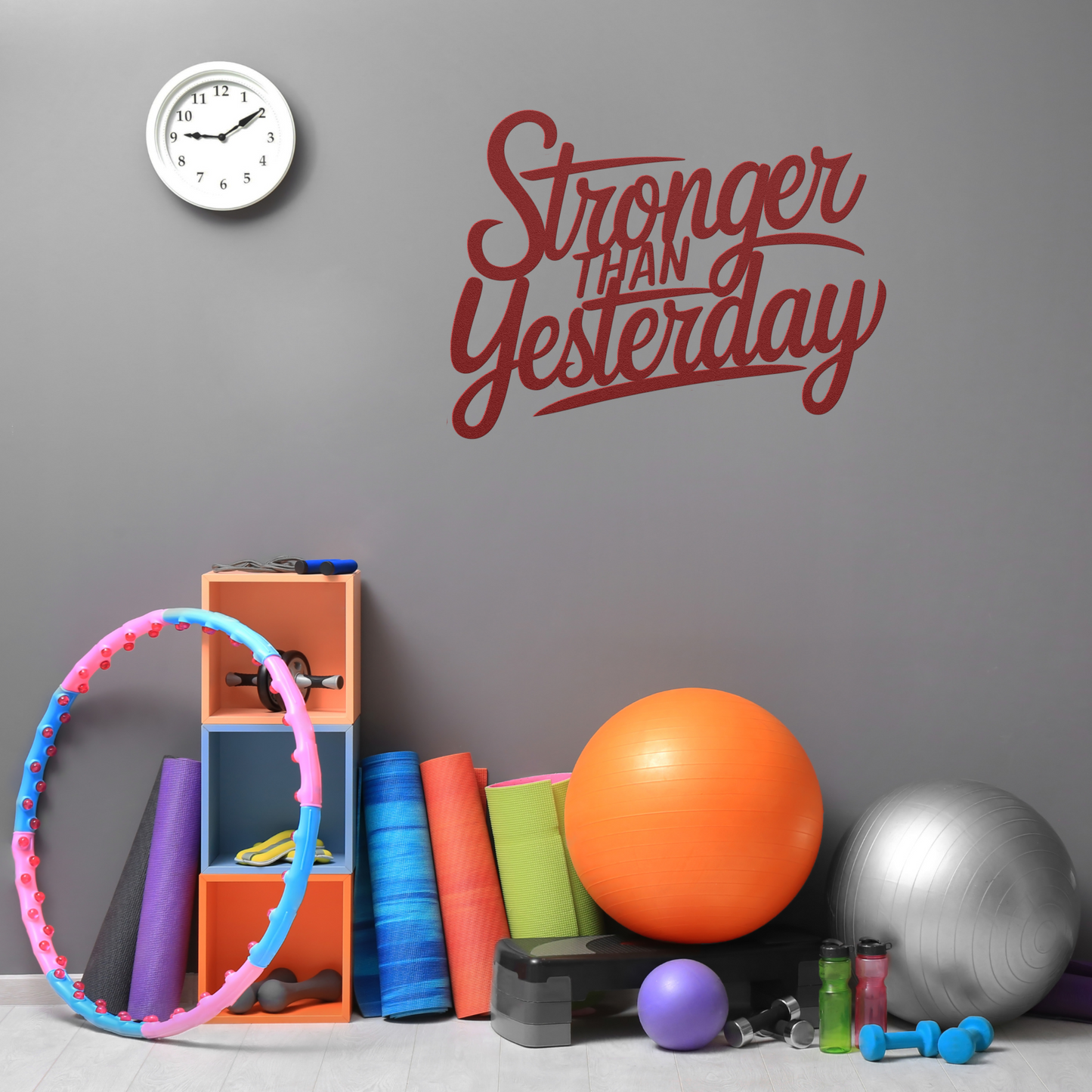In a gray gym, the "Stronger Than Yesterday Metal Sign" in vibrant red is displayed. Below it are powder-coated exercise mats, a hula hoop, stacked blocks, a kettlebell, an orange ball, and a gray stability ball. A wall clock reads 11:50.