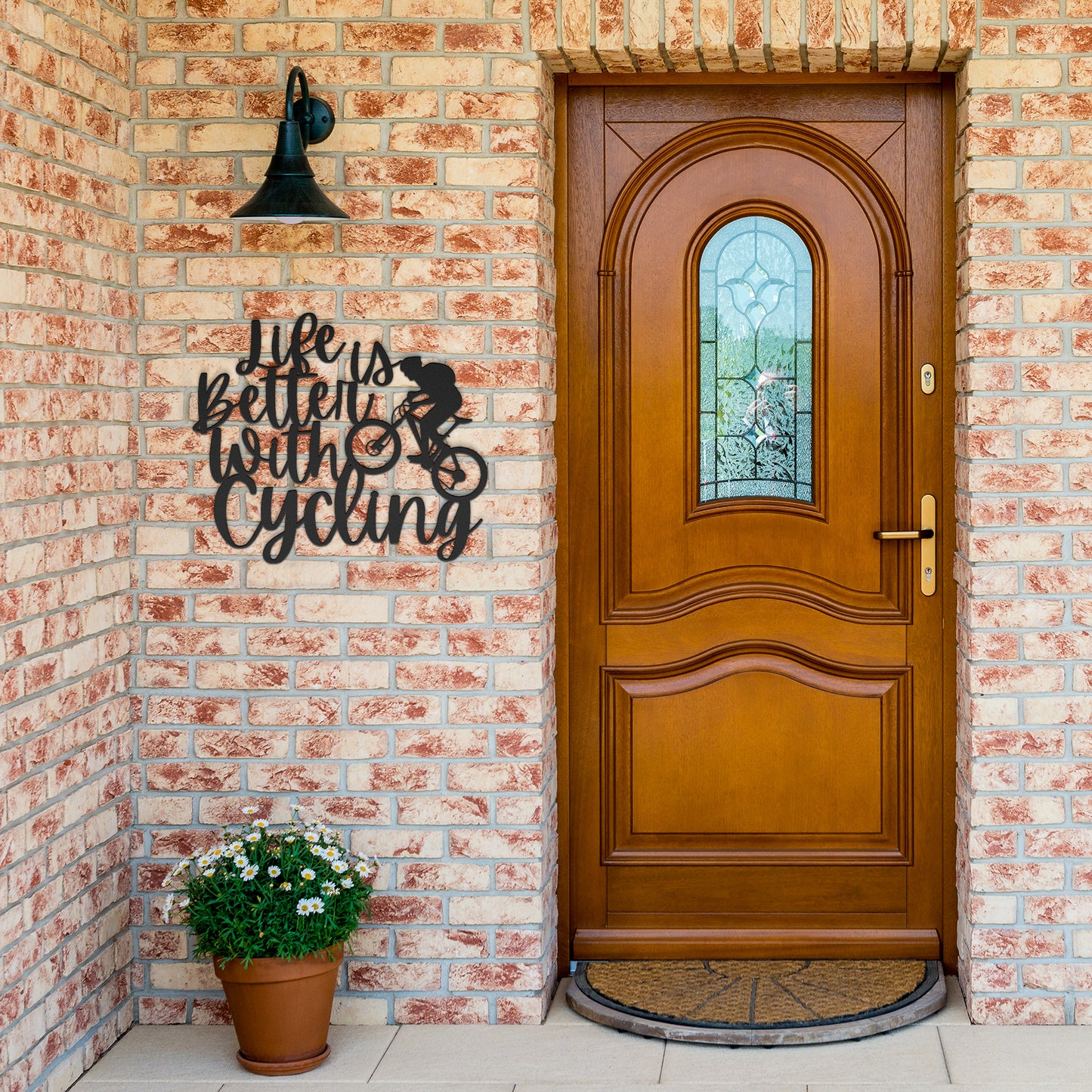 A brick wall with a wooden door, a durable Life Is Better With Cycling Metal Art sign, an exterior light, and a small potted plant with white flowers.