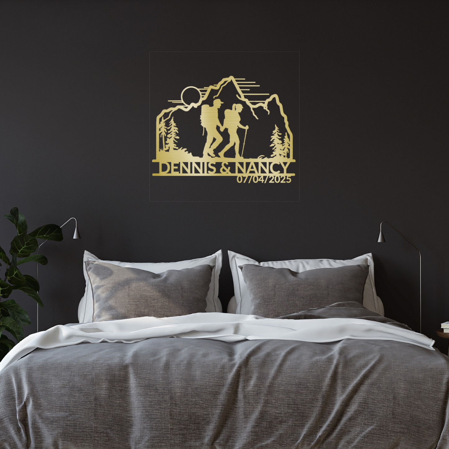 A modern bedroom showcases a double bed with gray bedding, white pillows, and the Personalized Hiking Couple Metal Wall Art above the bed, depicting a hiking couple, mountains, trees, and "DENNIS & NANCY" with the date 07/04/2025.