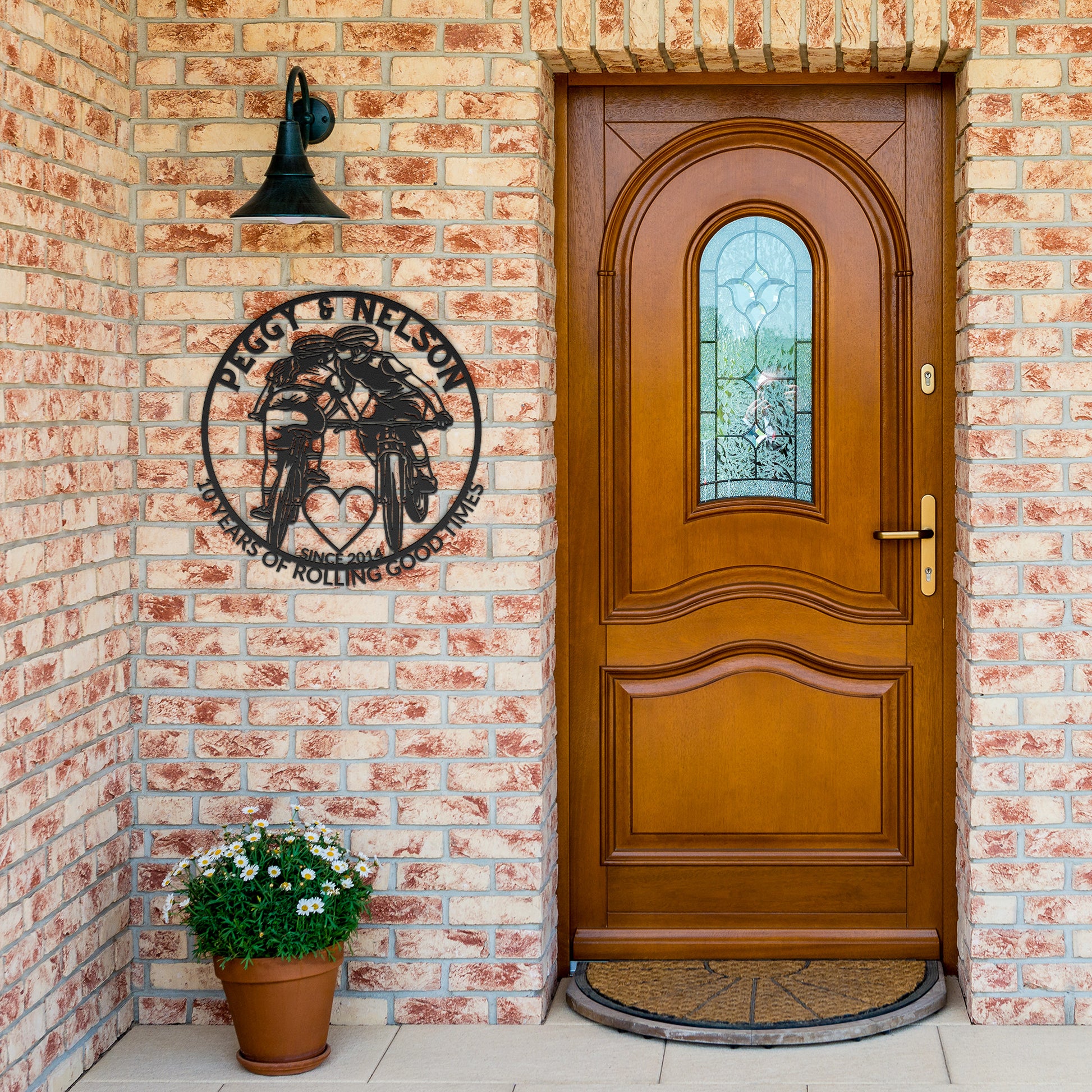 A wooden front door with a decorative glass panel is surrounded by brick walls. Above the doorbell hangs a custom metal art piece titled "Personalized Cycling Couple on Mountain Bikes," which reads "Peggy & Nelson, Masters of Rolling Goodness." Potted flowers adorn the porch, accentuating this unique gift for the cycling couple.