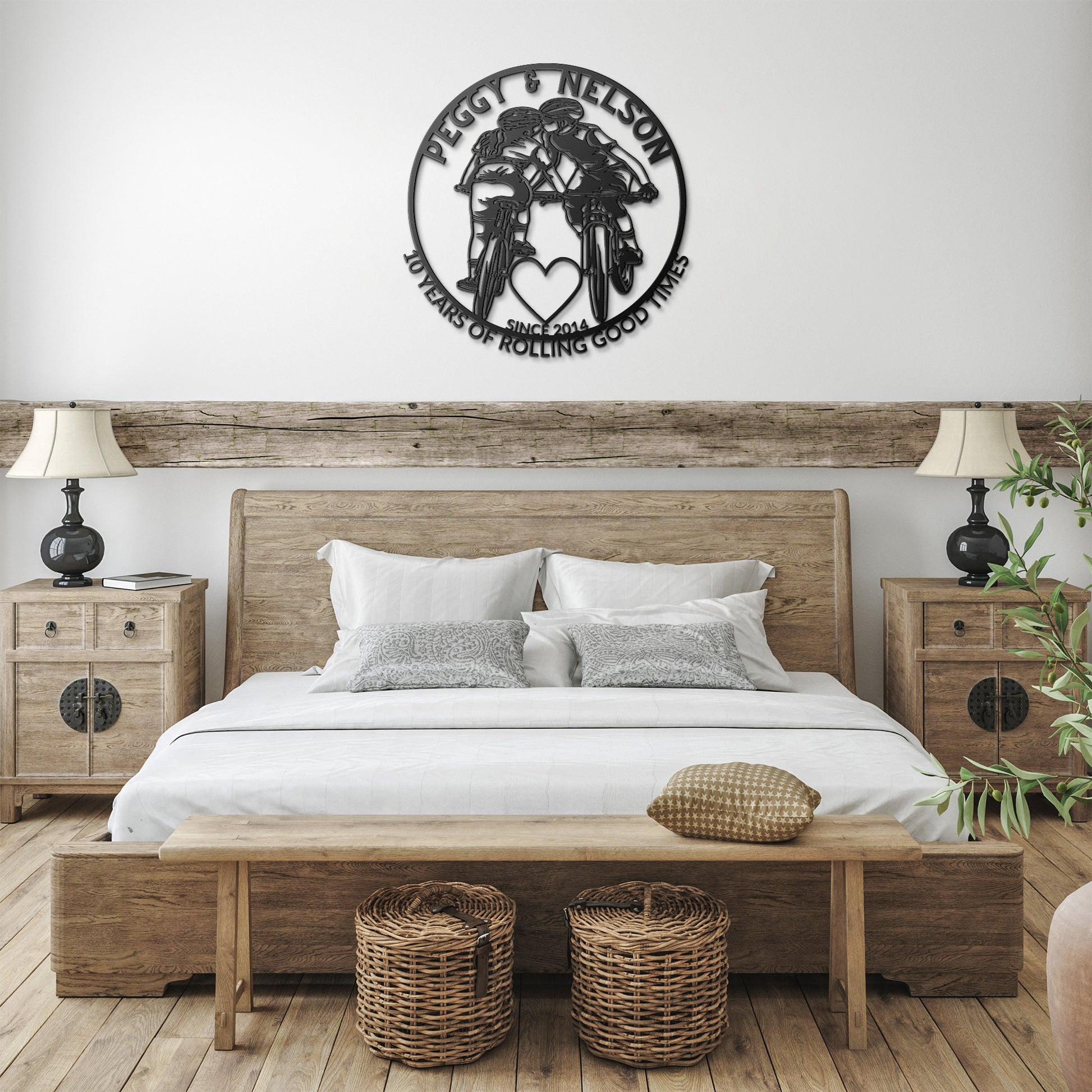 A cozy bedroom featuring a wooden bed, matching nightstands, and a bench. Two woven baskets are tucked underneath. Above the bed hangs a Personalized Cycling Couple on Mountain Bikes Metal Art Sign that reads "Peggy & Nelson, 20 Years of Rolling Good Times, Since 2004.
