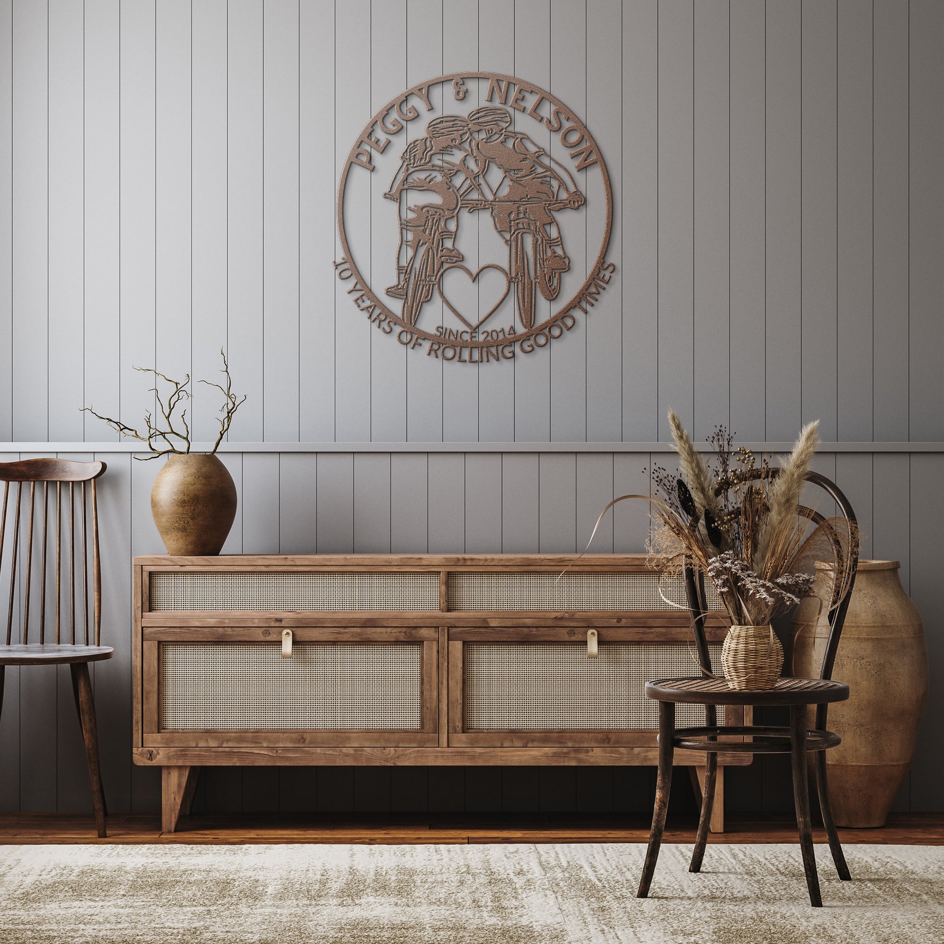 In a rustic room, a wooden chair, sideboard, and large vase create an inviting atmosphere. A personalized Cycling Couple on Mountain Bikes Metal Art sign reads "Peggy & Nelson, 10 Years of Rolling Good Times, Since 2014." Dried plants are elegantly displayed in vases.