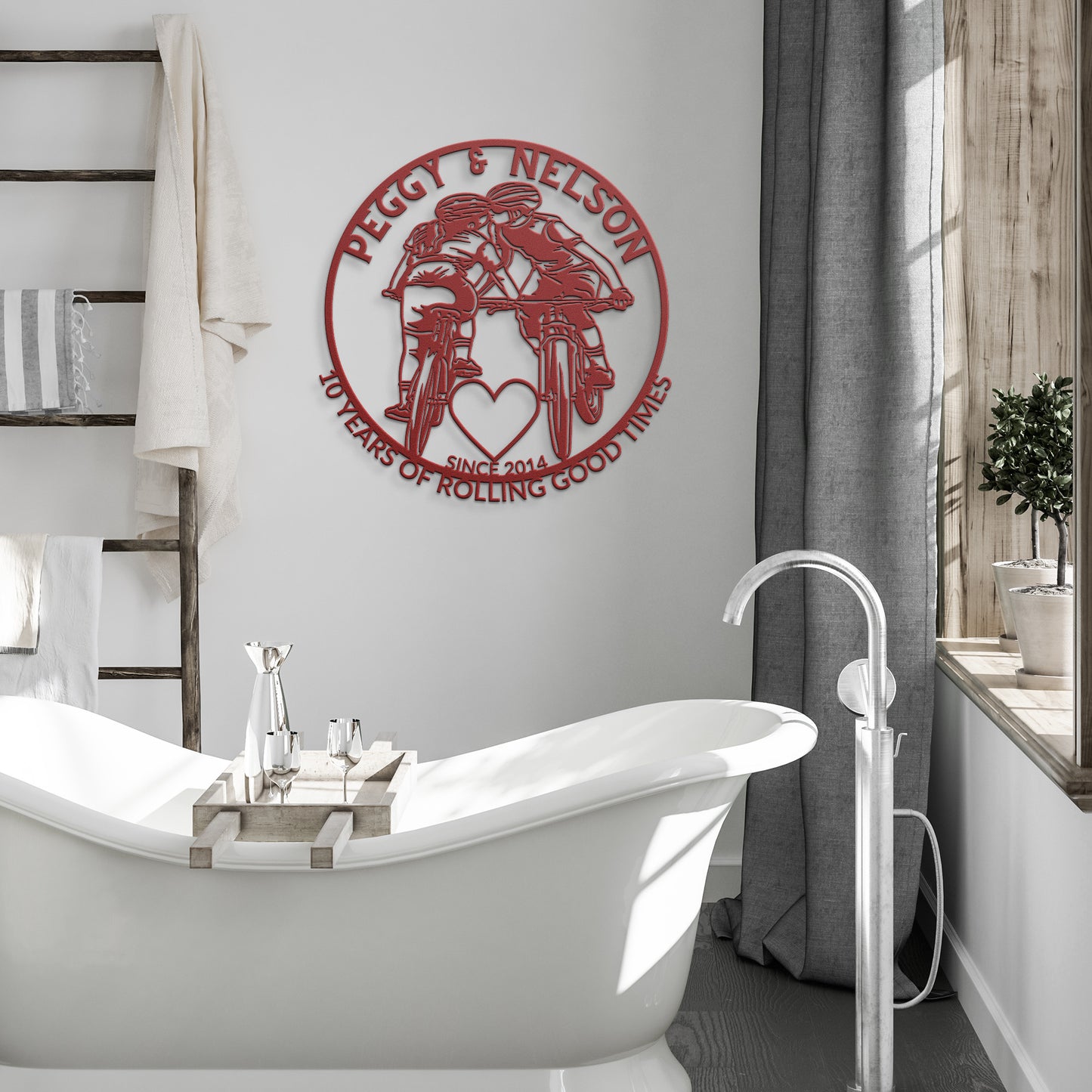 A charming bathroom features a freestanding tub, a shelf lined with towels, and a personalized cycling couple metal art sign depicting mountain bikes with hearts. The red metal sign reads: "Peggy & Nelson, 10 Years of Rolling Good Times, Since 2014," making it perfect as a gift for a cycling couple.