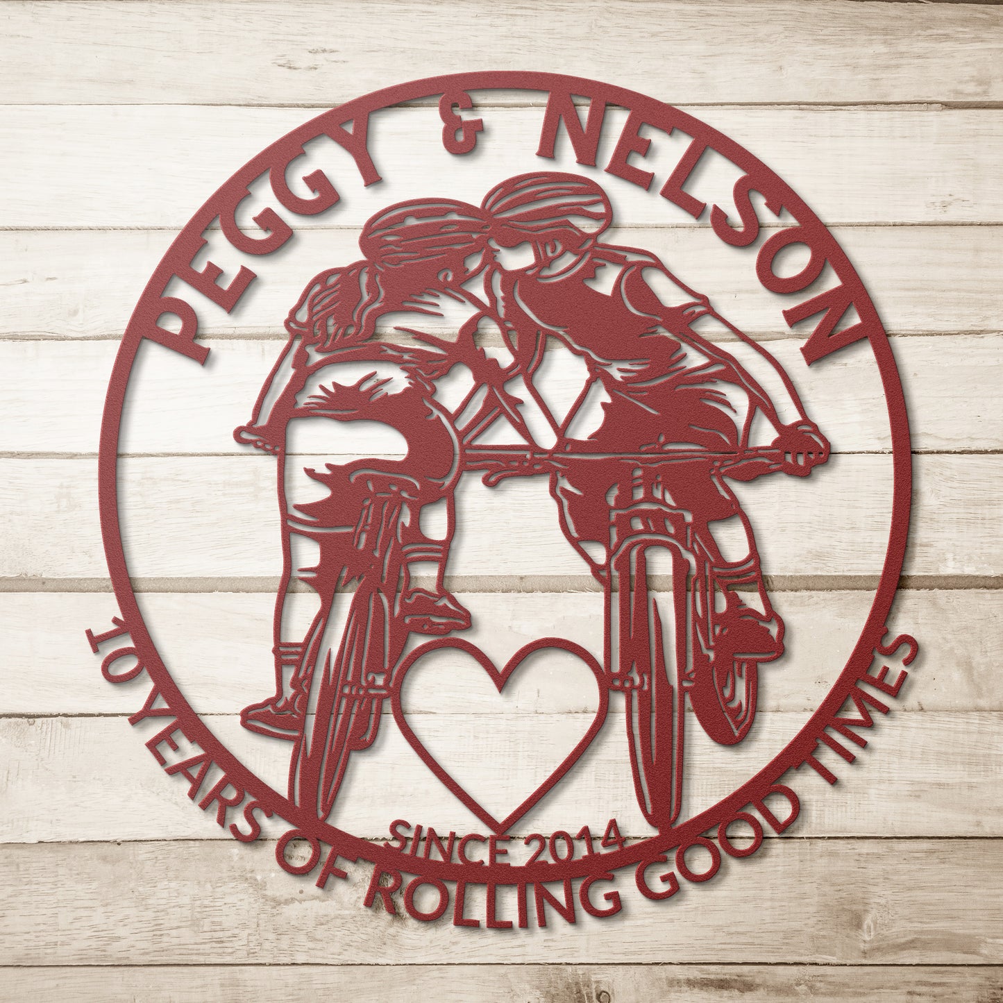 A personalized metal art sign featuring a silhouette of two cyclists kissing inside a red circle is set against a wooden background. It includes the text "Peggy & Nelson, 10 Years of Rolling Good Times, Since 2014." The Personalized Cycling Couple on Mountain Bikes Metal Art is the perfect gift for bicycle enthusiasts to celebrate love and adventure.