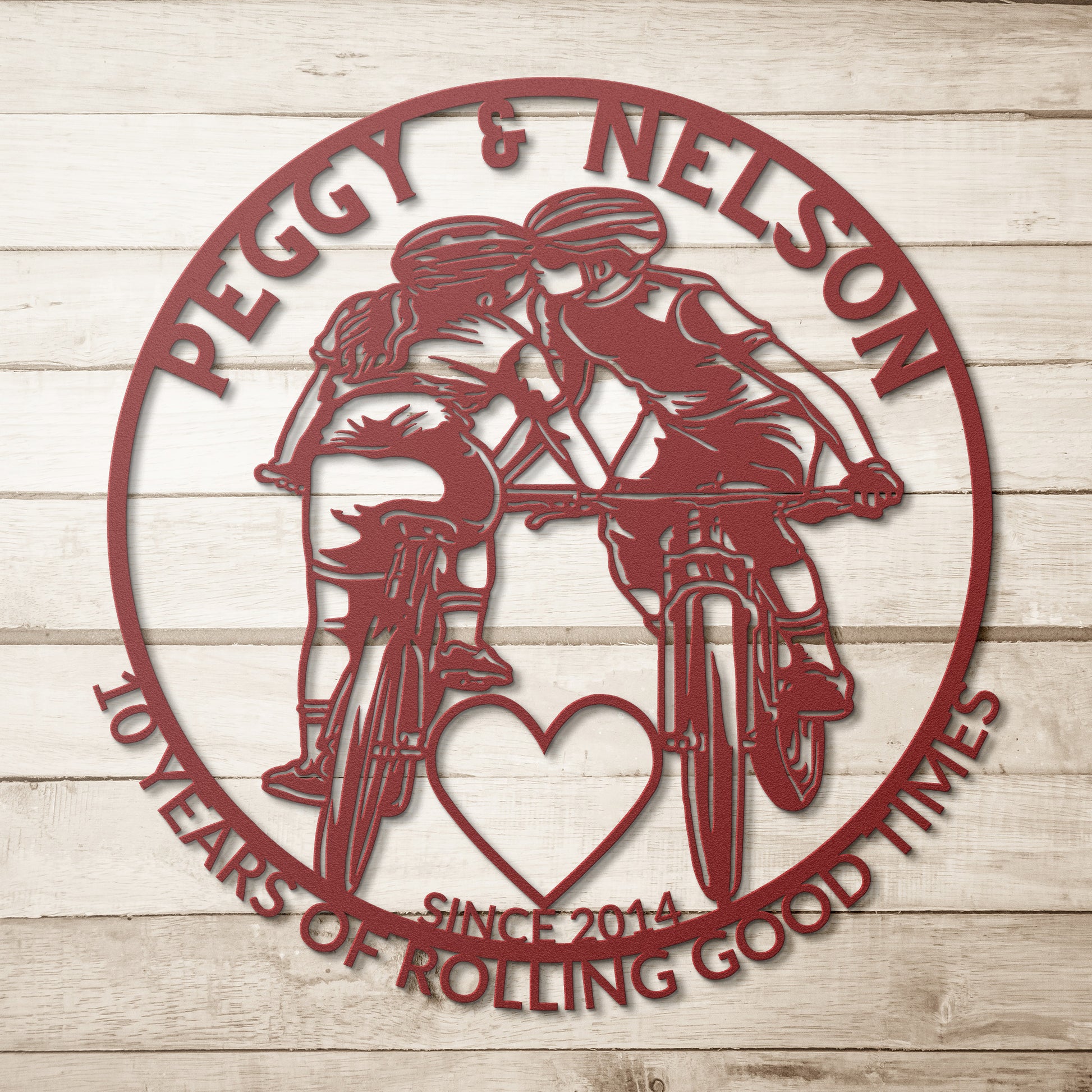 A personalized metal art sign featuring a silhouette of two cyclists kissing inside a red circle is set against a wooden background. It includes the text "Peggy & Nelson, 10 Years of Rolling Good Times, Since 2014." The Personalized Cycling Couple on Mountain Bikes Metal Art is the perfect gift for bicycle enthusiasts to celebrate love and adventure.