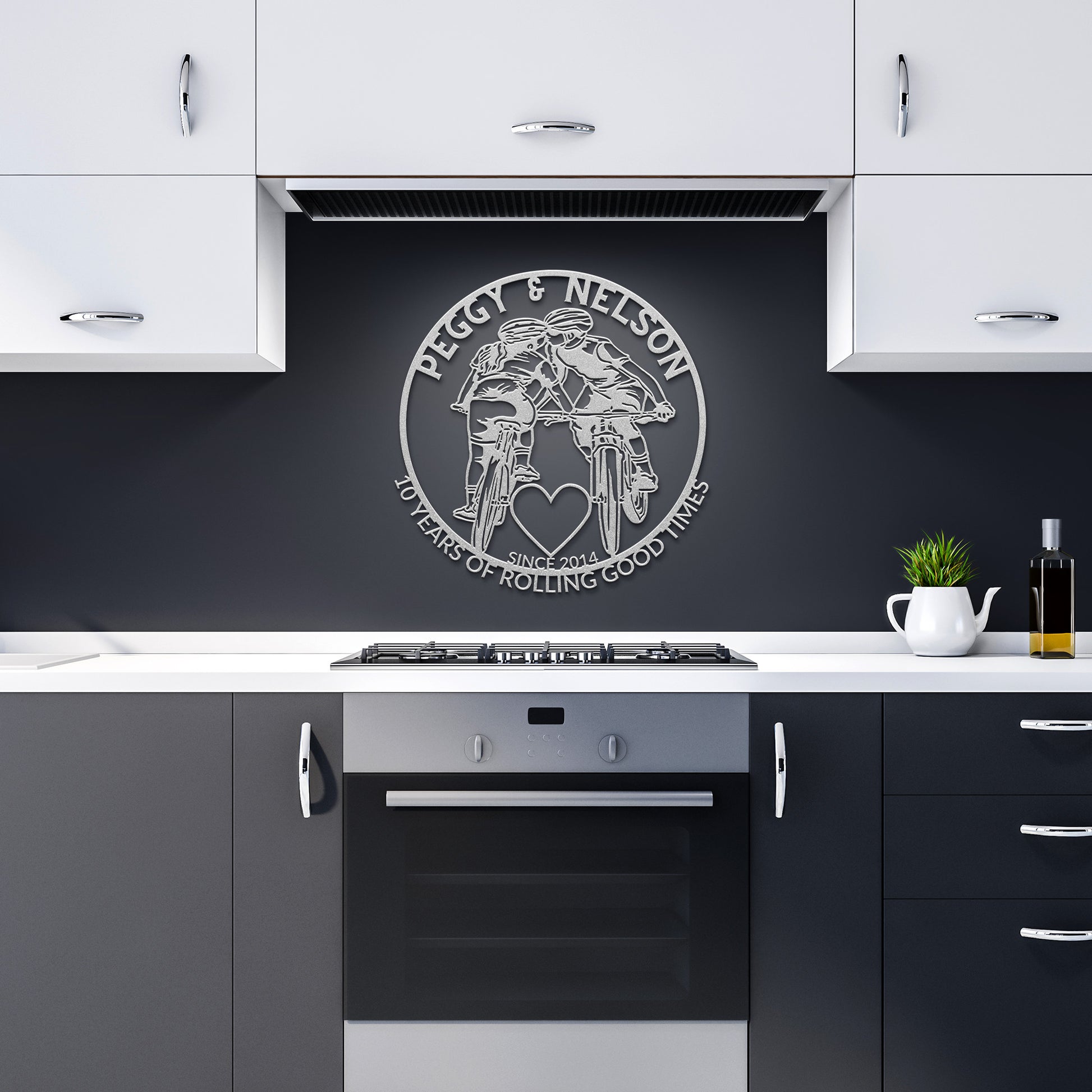 A kitchen with black and white cabinets showcases a wall decoration depicting two motorcyclists, featuring the text "Peggy & Nelson: 40 Years of Rolling Good Times," and a heart above an oven—an ideal version of the Personalized Cycling Couple on Mountain Bikes Metal Art for those who cherish their journey together.