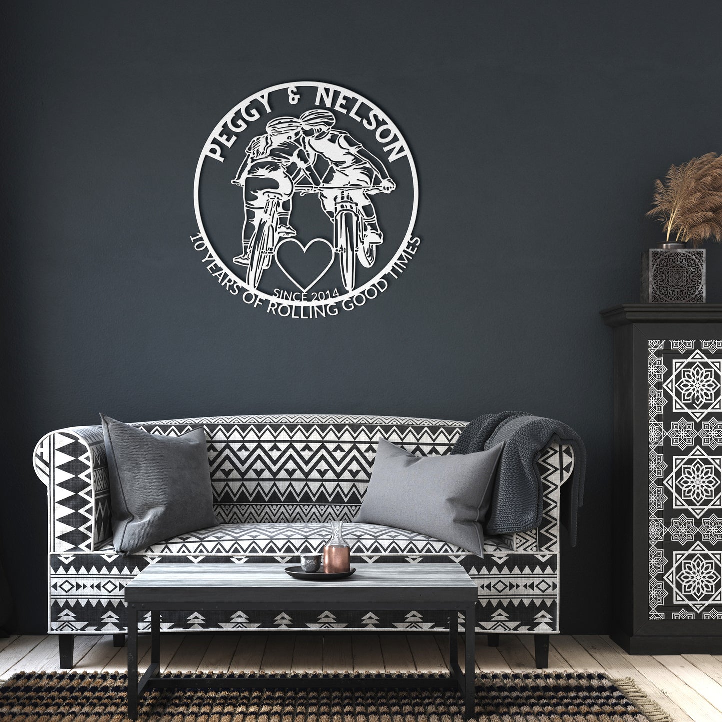 A black and white living room showcases a geometric-patterned sofa, cushions, and a Personalized Cycling Couple on Mountain Bikes Metal Art. The plaque features two cyclists and reads, "Peggy & Nelson: 10 Years of Rolling Good Times, Since 2014.