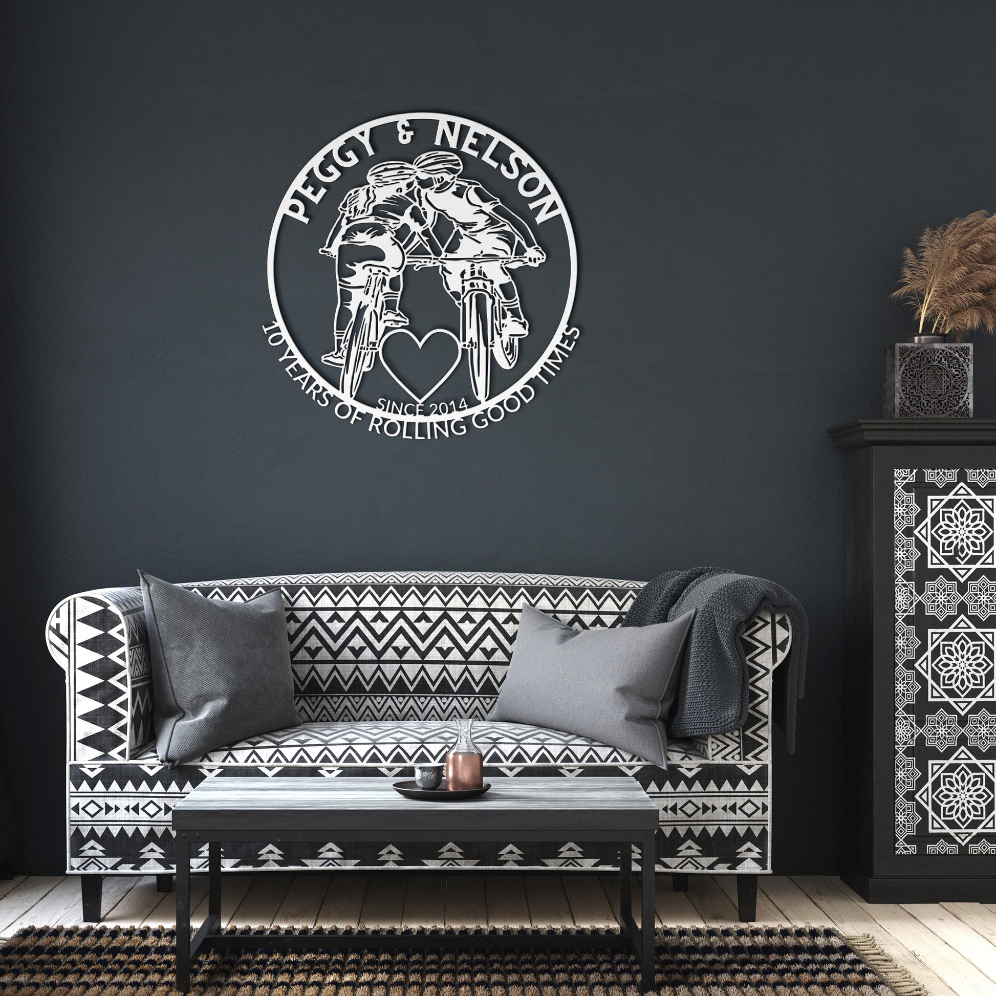 A black and white living room showcases a geometric-patterned sofa, cushions, and a Personalized Cycling Couple on Mountain Bikes Metal Art. The plaque features two cyclists and reads, "Peggy & Nelson: 10 Years of Rolling Good Times, Since 2014.