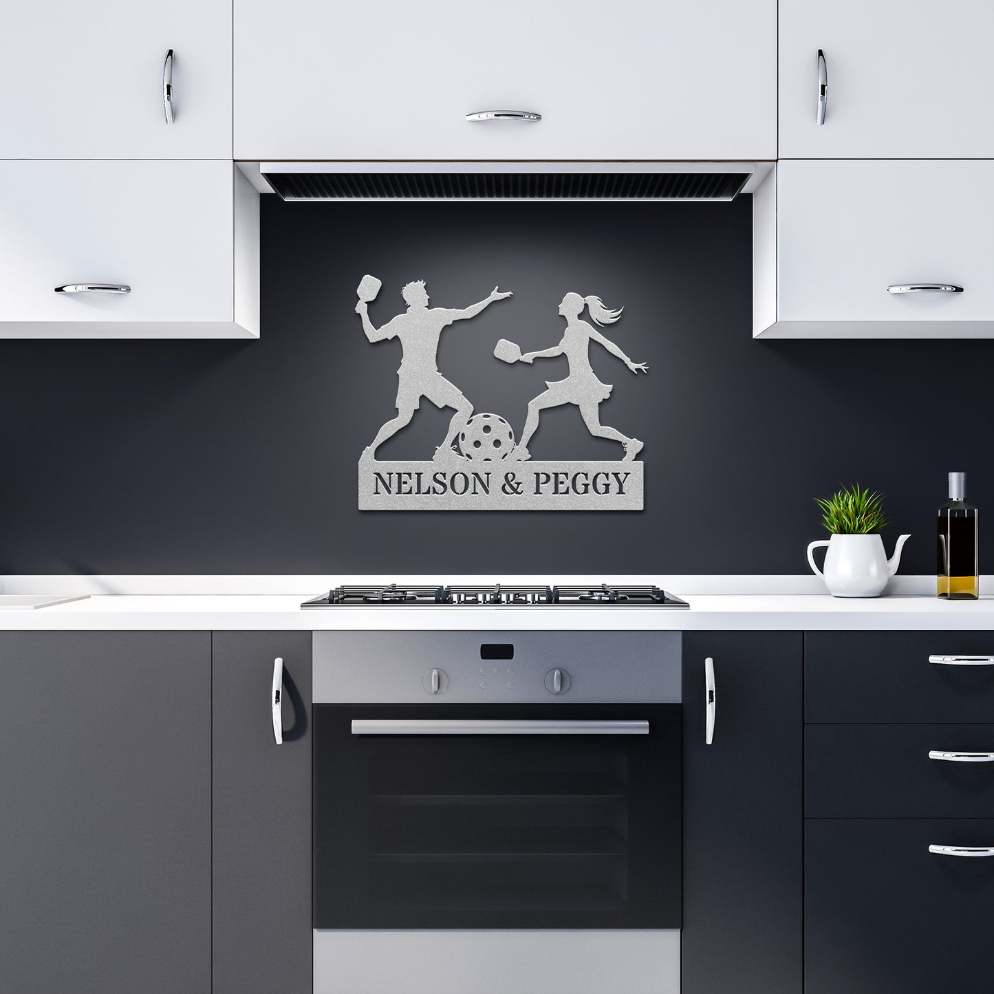 In the modern kitchen with white and gray cabinets, a stylish metal wall art crafted from 18-gauge steel highlights a couple playing sports. A personalized Pickleball Couple Names Metal Sign labeled "Nelson & Peggy" is prominently displayed above the stovetop, adding a unique touch to the home decor.