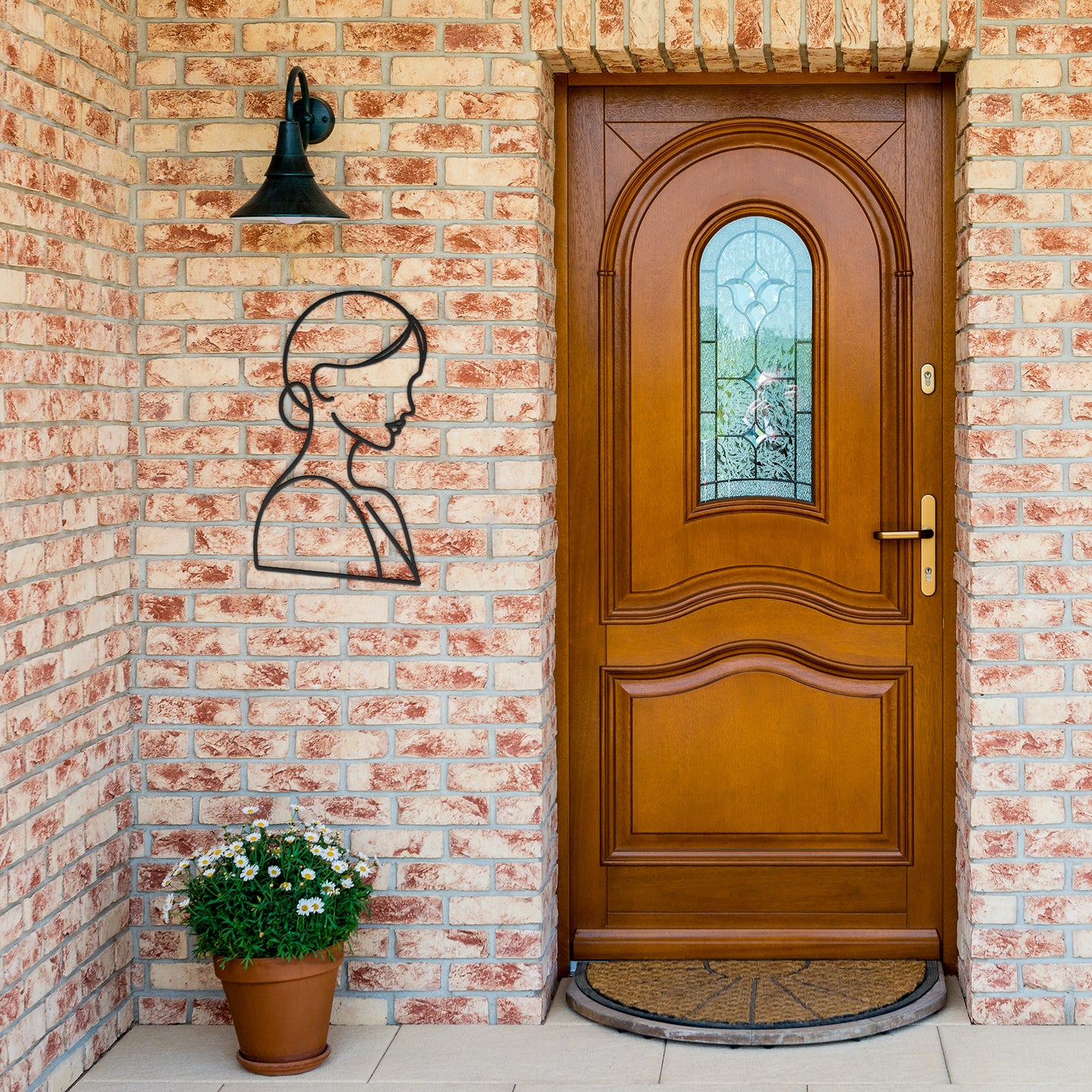 A wooden arched front door with a stained glass window set in a brick wall, adorned with the Woman Line Art powder-coated metal wall silhouette and a potted plant with white flowers. A black outdoor light fixture is above the art.