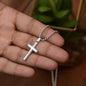 A ShineOn Fulfillment Prayer For Graduation - Cross Necklace with Ball Chain rests in the palm of a hand with a plant blurred in the background.