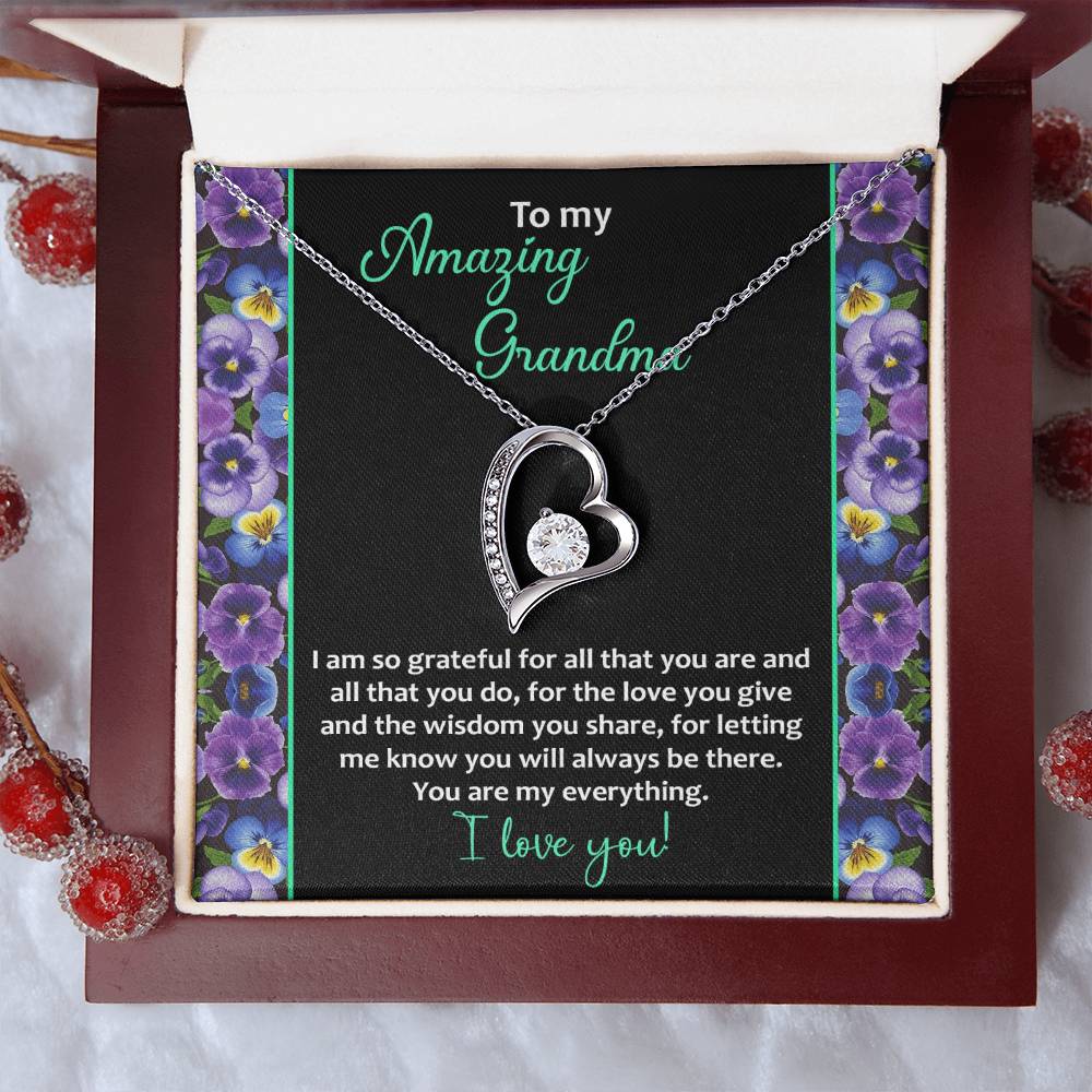 The "To Grandma, My Everything - Forever Love Necklace" is beautifully displayed in an open box. The heart-shaped pendant, showcasing a small inner heart, exudes timeless elegance with its gleaming 14k white gold finish and sparkling cubic zirconia stones. The box features a heartfelt message for a grandmother on a black card embellished with purple flowers and green text.