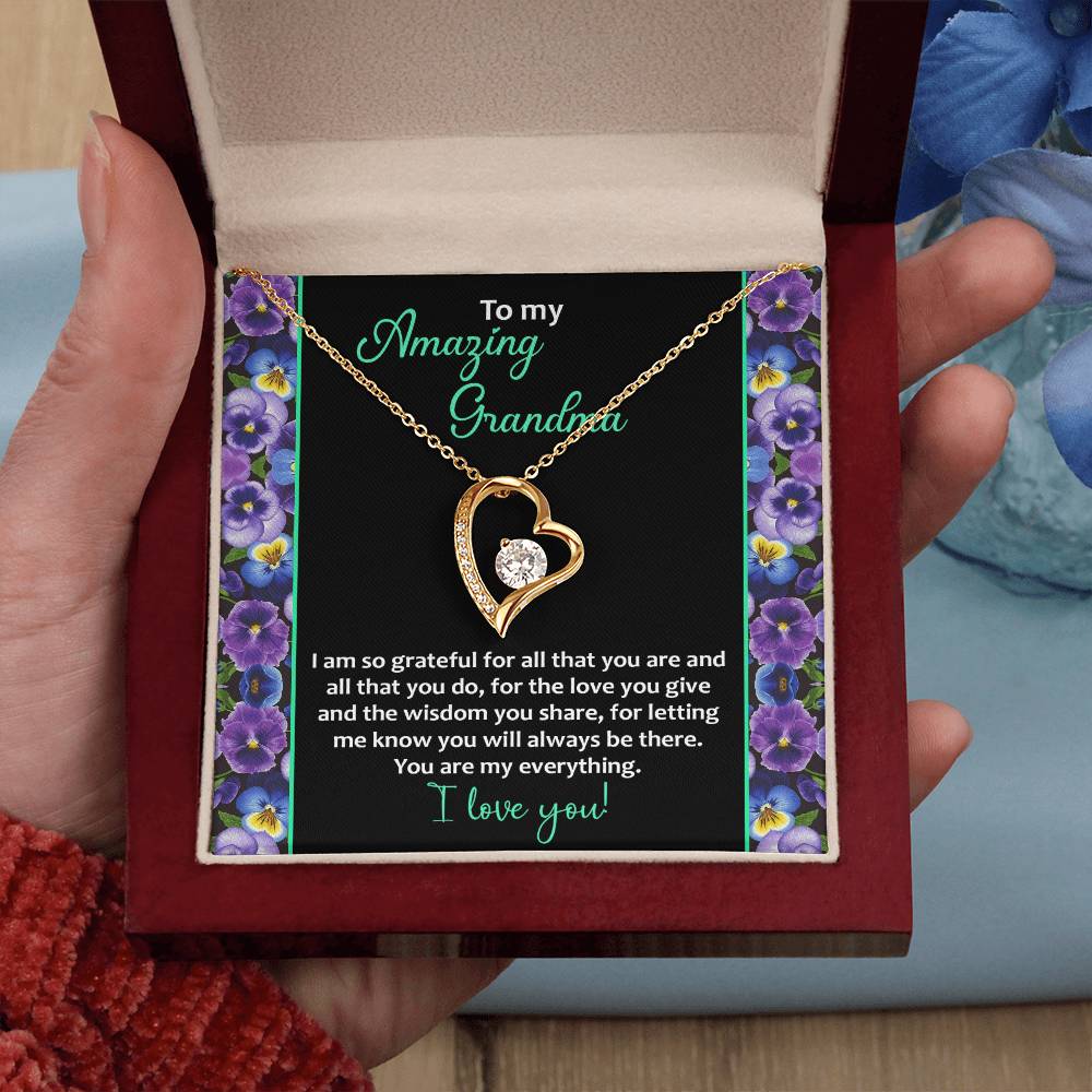 A hand holds a jewelry box containing the "To Grandma, My Everything - Forever Love Necklace," featuring a heart-shaped pendant with a cubic zirconia stone. The inner lid of the box is adorned with a floral border and bears a heartfelt message expressing gratitude and love for a grandmother, encapsulating the timeless beauty of the necklace.