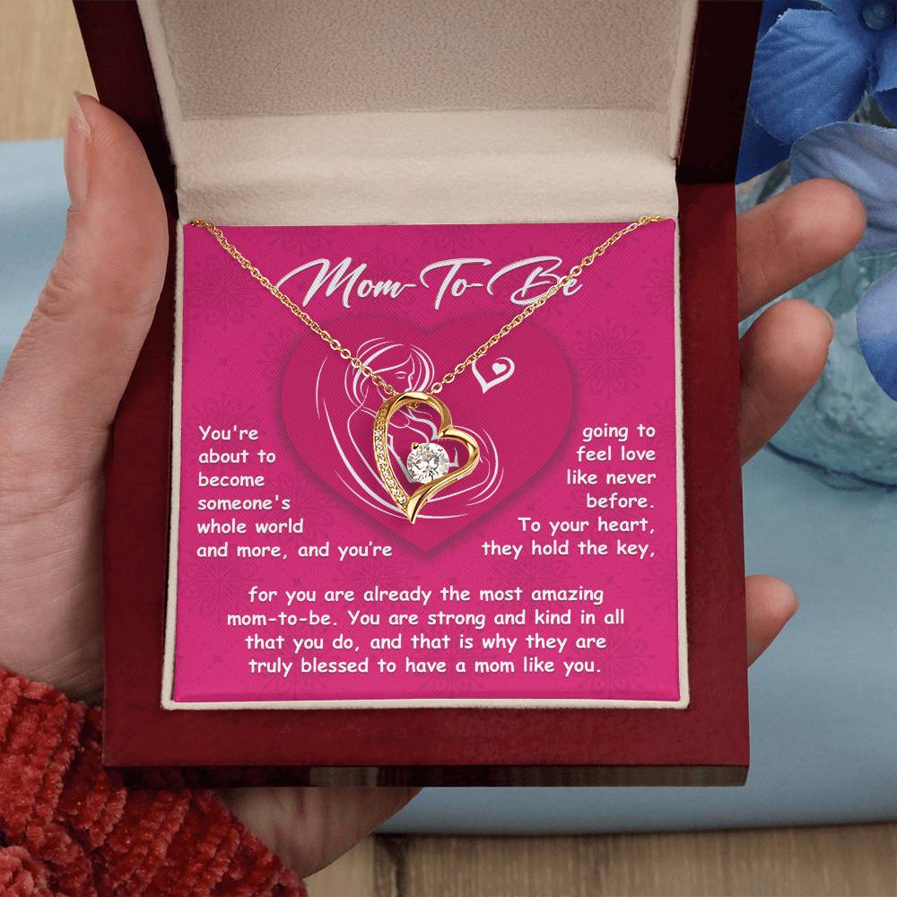 A hand holds a jewelry box containing the "To Mom To Be, Someone's Whole World - Forever Love Necklace," which features an exquisite heart and knot design. Behind the necklace, there is a card titled "Mom-To-Be" showcasing a heartfelt message. Blue flowers in the background enhance its charm, making it an ideal choice for personalized gifts.