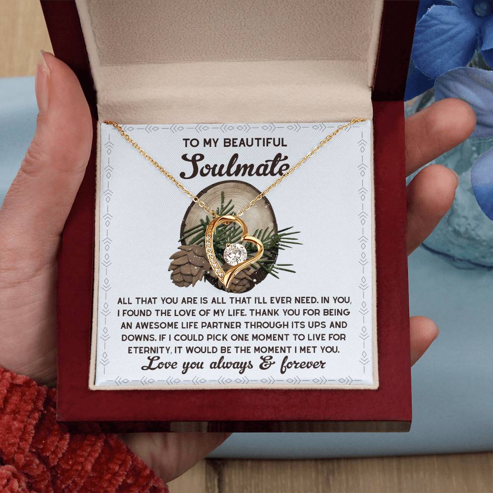 A hand holds a wooden box containing the Soulmate-One Moment - Forever Love Necklace. This necklace, featuring intertwined hearts and adorned with CZ crystals, is accompanied by a loving message about partnership on the inside lid. You can choose between an elegant white gold or classic yellow gold finish to complete this timeless piece.