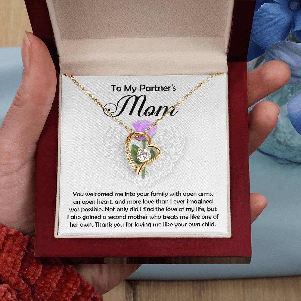 A hand holds an open jewelry box with the Partner's Mom-A Second Mother - Forever Love Necklace, featuring a gold-finished cubic zirconia heart pendant and a heartfelt message insert for a partner’s mother. Blue flowers are nearby.