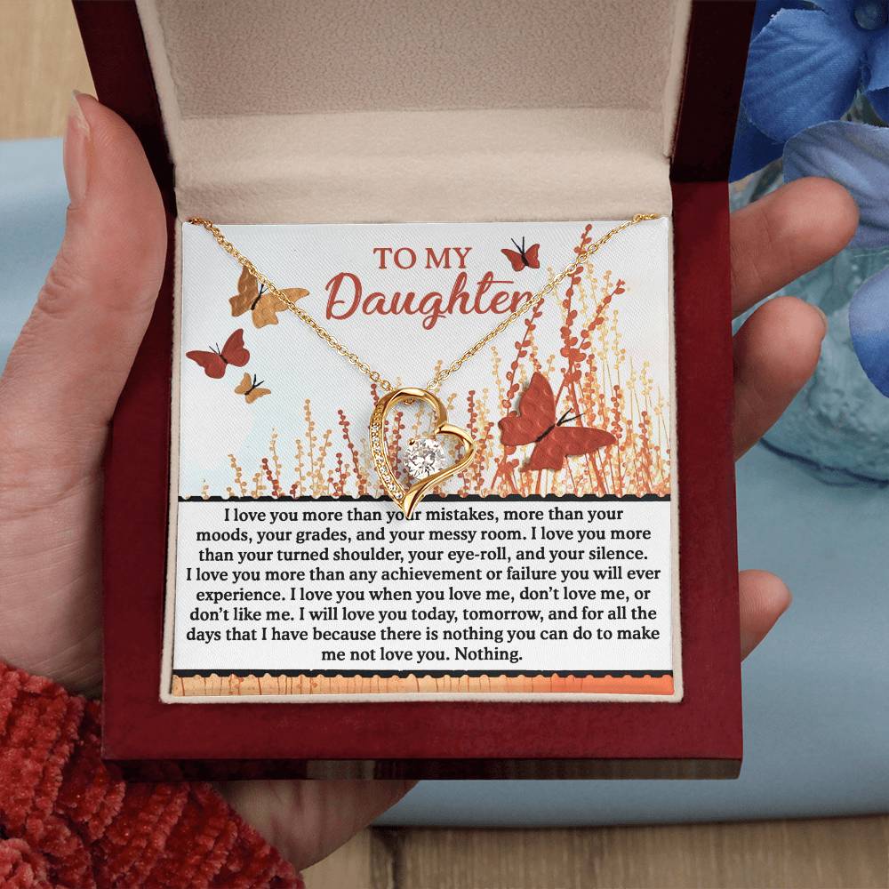 A hand presents an open box showcasing the Daughter-Love You More - Forever Love Necklace, featuring a heart pendant embellished with sparkling CZ crystal. The inside of the box reveals a touching message that says "To My Daughter," surrounded by butterflies and flowers, all elegantly highlighted with a delicate gold finish.