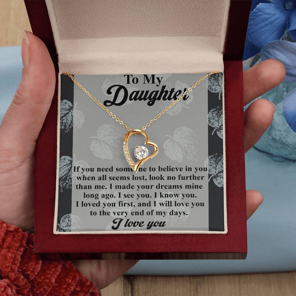 A hand holds a jewelry box containing the Daughter-My Days - Forever Love Necklace, featuring a heart-shaped design adorned with CZ crystals and finished in gleaming white gold. The message reads "To My Daughter," along with a heartfelt note inside.