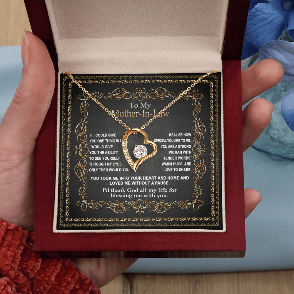 A hand holds an open red box containing the "To Mother-In-Law, Through My Eyes - Forever Love Necklace," featuring a heart-shaped pendant. Inside the box is a little message addressed to your mother-in-law.