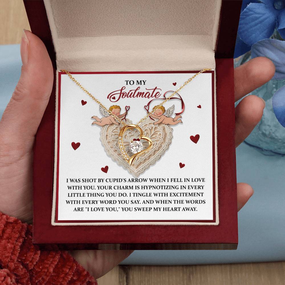A person holds an open box displaying the Soulmate-Sweep My Heart - Forever Love Necklace, featuring a heart pendant adorned with CZ crystals and a gold finish. It rests on a card with two cupids and an enchanting romantic message.
