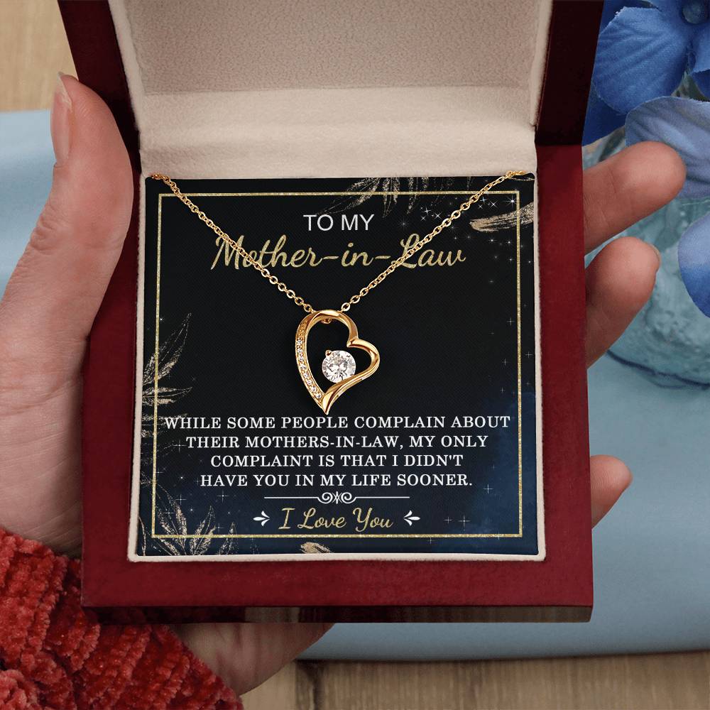 A person holds the "To Mother-In-Law, In My Life - Forever Love Necklace" with a heart-shaped pendant in an open gift box. The pendant, embellished with a sparkling cubic zirconia stone and set in a gold finish, is accompanied by a message that reads, "To my Mother-in-law...my only complaint is that I didn't have you in my life sooner.
