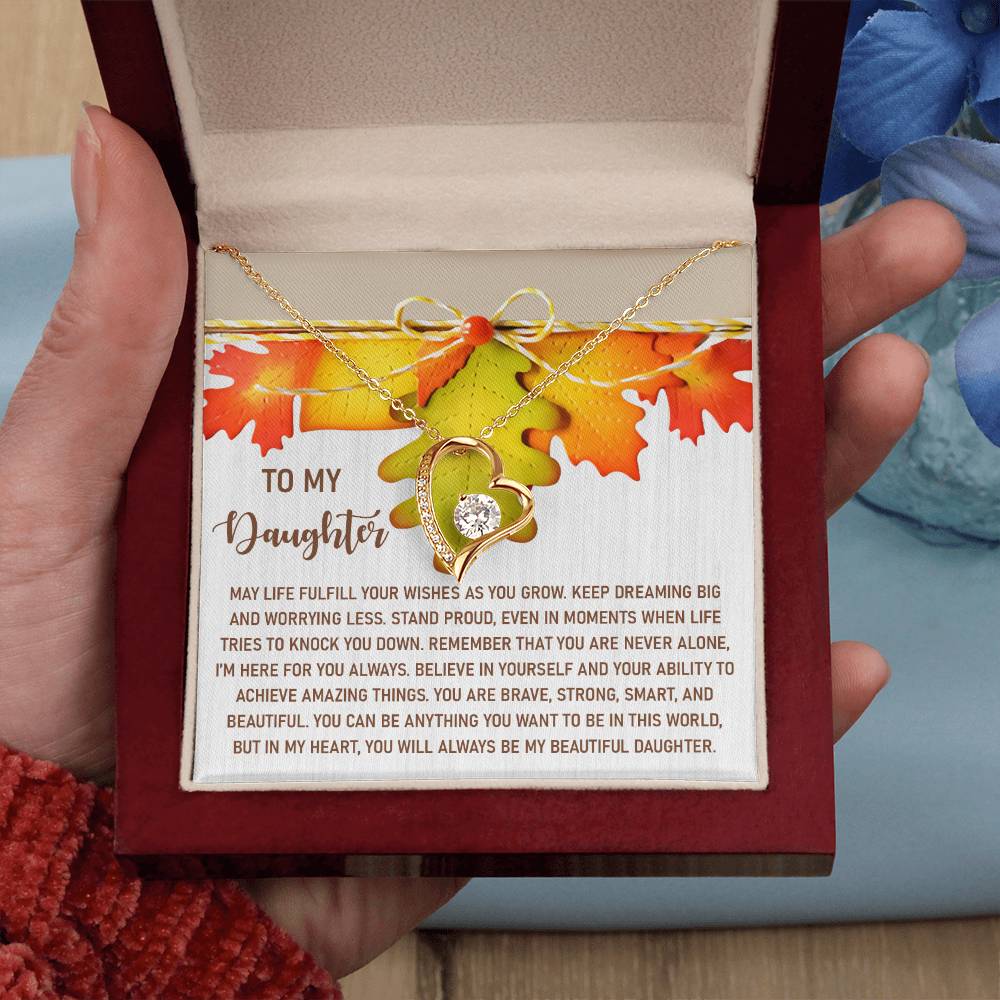 A person holds a decorative box with a gold finish containing the Daughter-For You Always - Forever Love Necklace and a heartfelt message titled "To My Daughter," adorned with shimmering cubic zirconia and surrounded by autumn leaves.