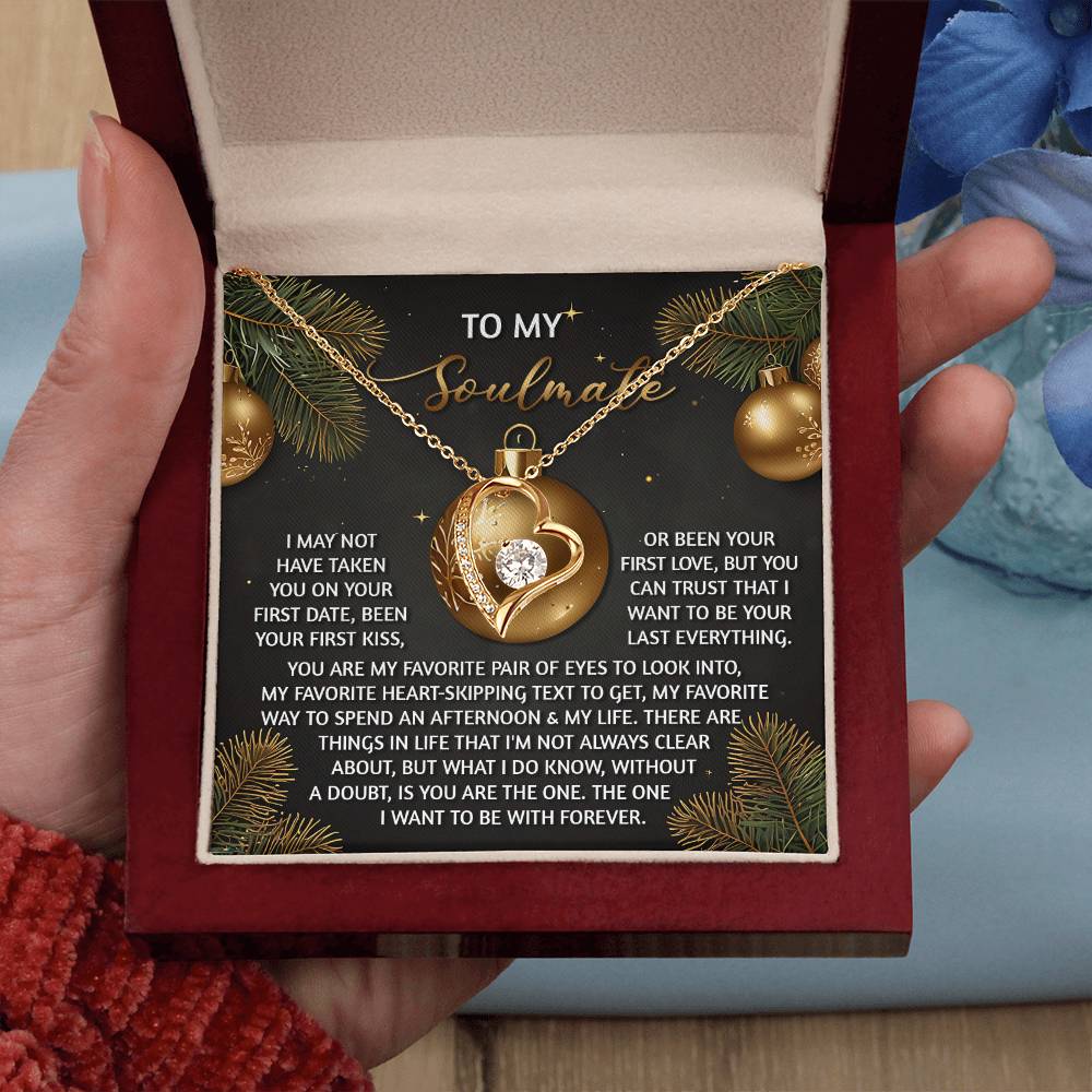 A hand holds a box containing the "Soulmate-Without A Doubt - Forever Love Necklace," featuring a heart-shaped pendant with a small gem. In the background, decorative holiday ornaments create the perfect setting for this symbol of eternal love.