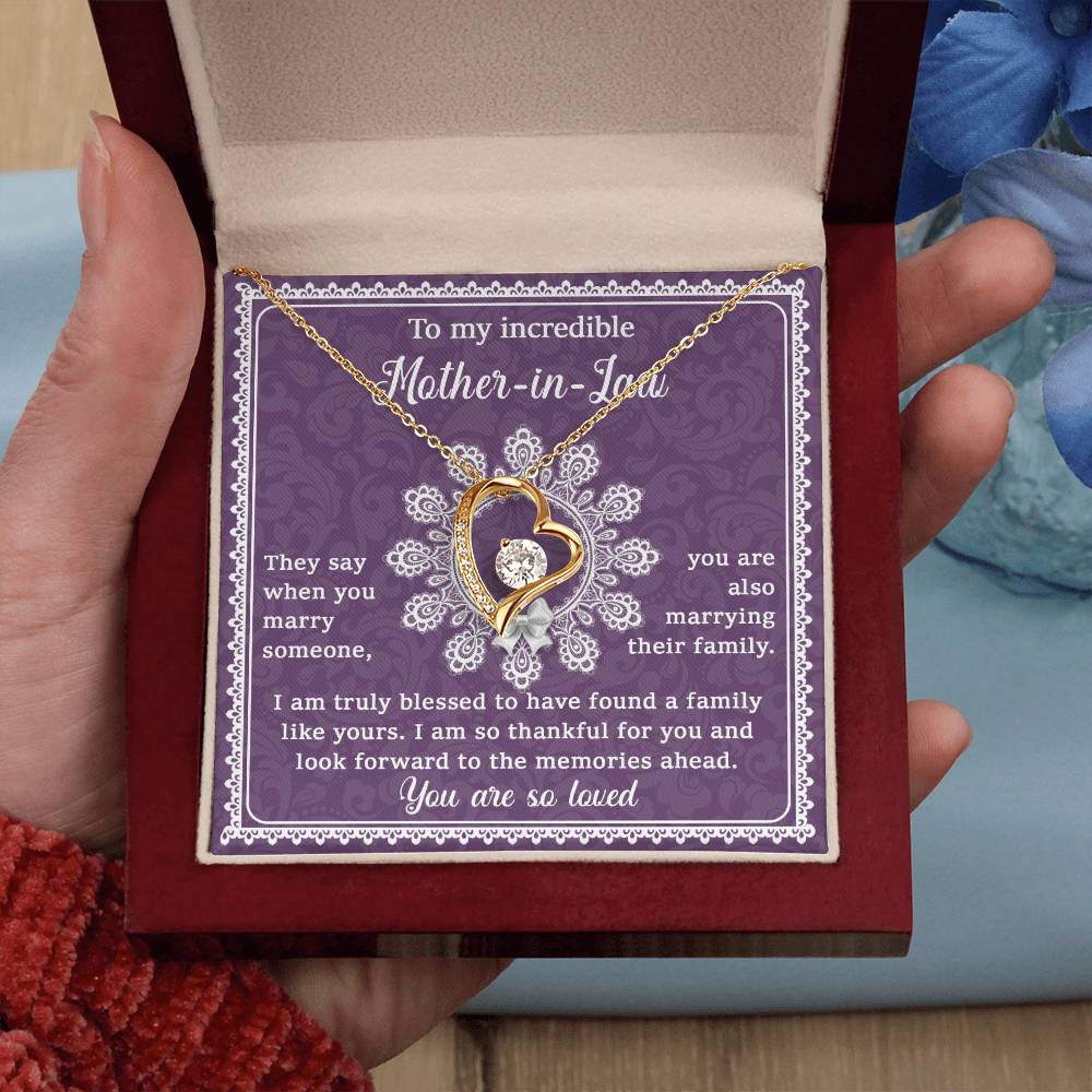 A hand holding an open jewelry box reveals the "To Mother-In-Law, Family Like Yours - Forever Love Necklace," featuring a heart-shaped pendant with a 14k white gold finish. The lid displays a heartfelt message dedicated to a mother-in-law, making it a unique gift. Blue flowers in the background highlight the elegance of this beautiful necklace.