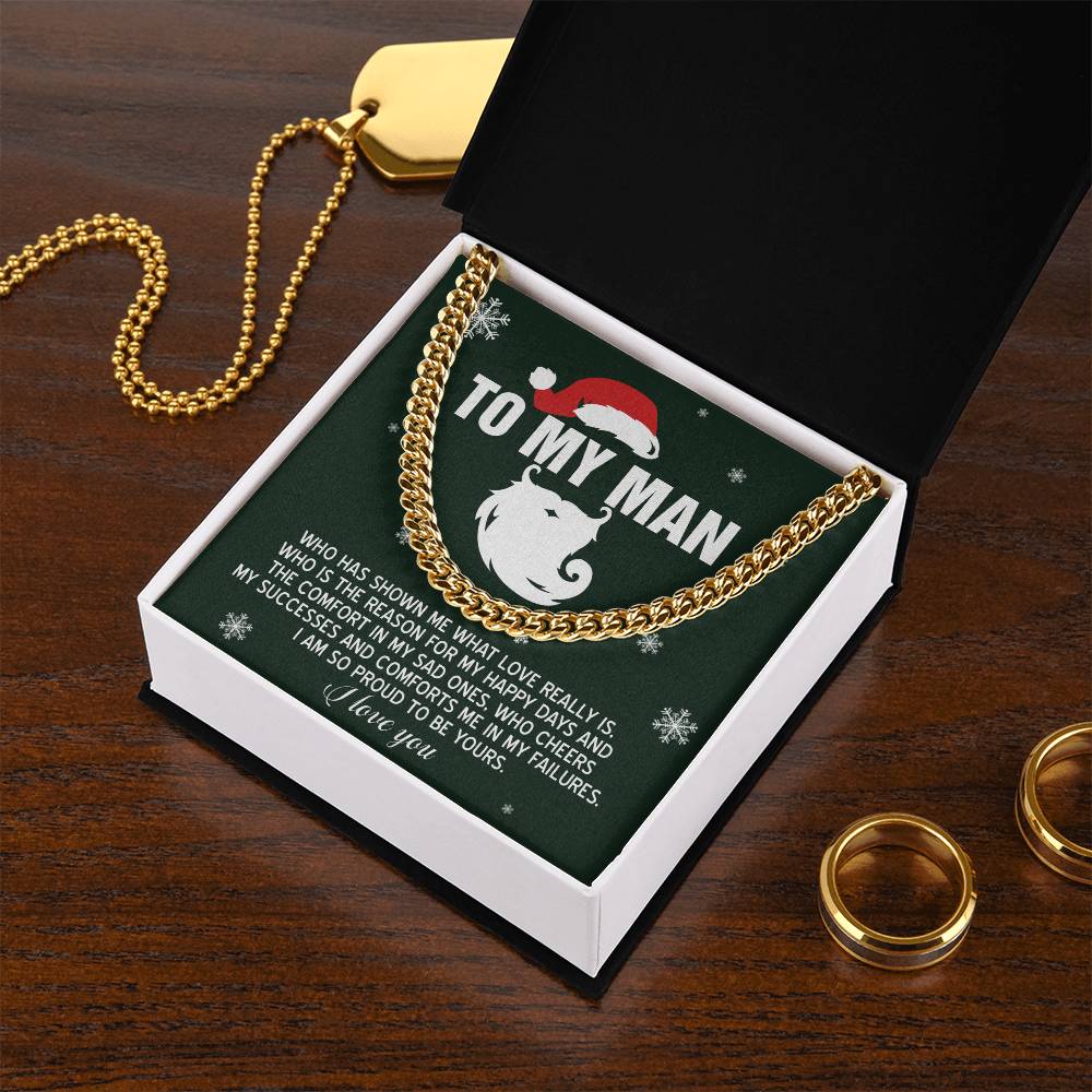 The "My Man-To Be Yours" Cuban Link Chain necklace, made of polished stainless steel in a gold finish, is presented on a green card within a black box. The card features the message "To My Man" with a Santa hat graphic, conveying feelings of love and pride. Accents like gold rings and a dog tag rest on the wooden surface, enhancing the sophistication of this elegant necklace design.