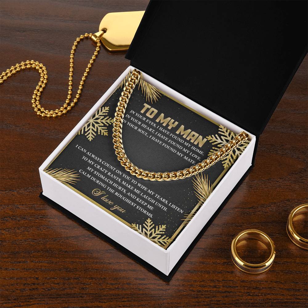 A Cuban Link Chain necklace named "My Man-Found My Home" is elegantly displayed in an open black box with a personalized message inscribed on the inner lid. Next to it, two gold rings and a gold dog tag are placed on a wooden table, creating the perfect setting for a thoughtful, personalized gift.