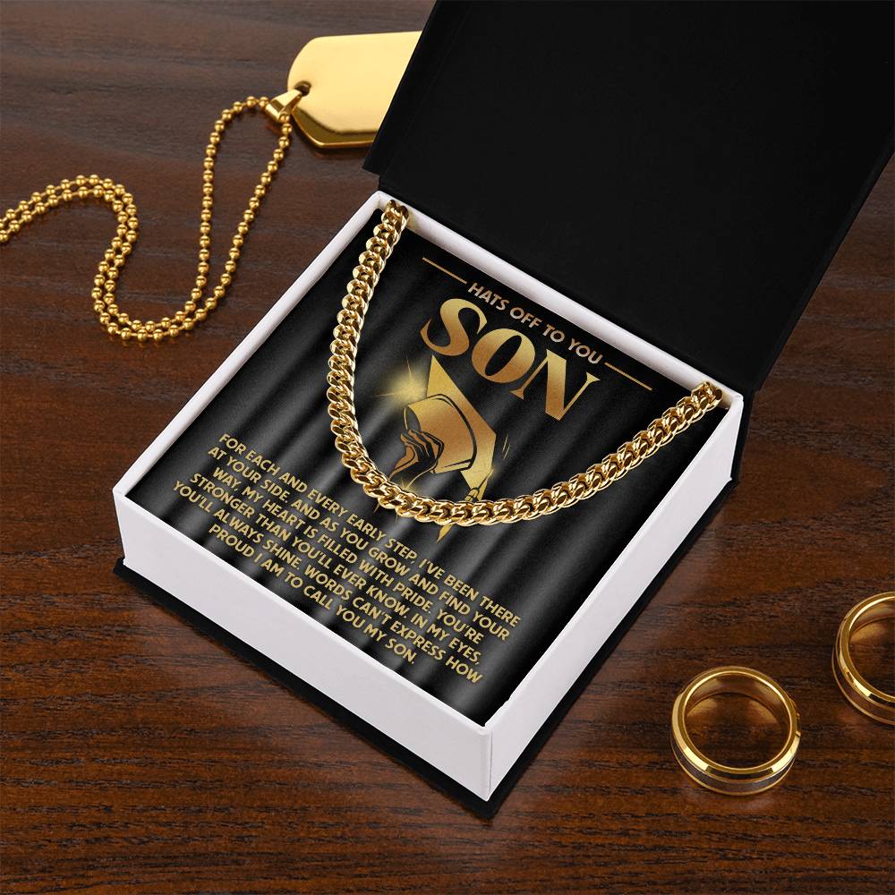 The "To Son, You'll Always Shine - Cuban Link Chain," a 14K yellow gold necklace, is showcased in an open black gift box with the message "Hats Off to You Son" inside. The box rests on wood, surrounded by rings and a gold dog tag.