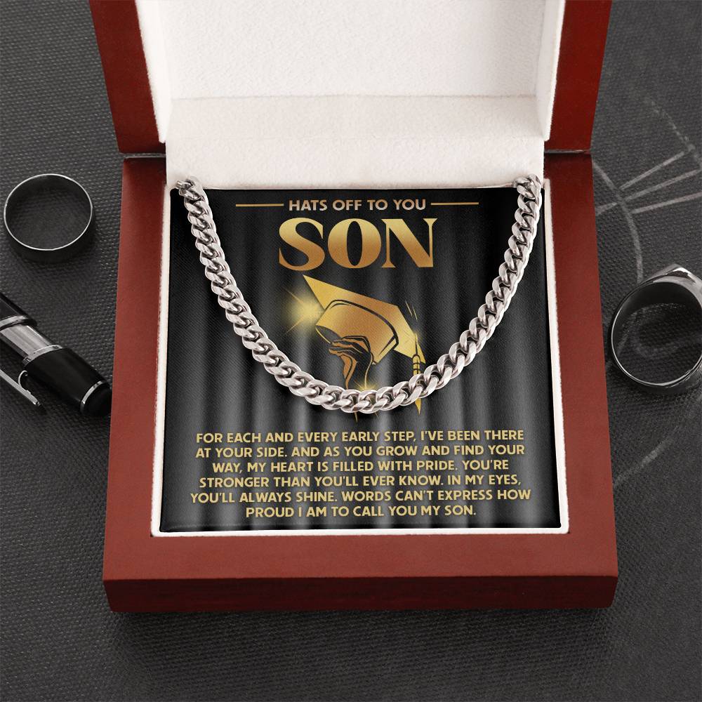 A 14K yellow gold Cuban link chain necklace, named "To Son, You'll Always Shine," rests in a red gift box with a heartfelt message. Rings and a pen are nearby on the dark surface.