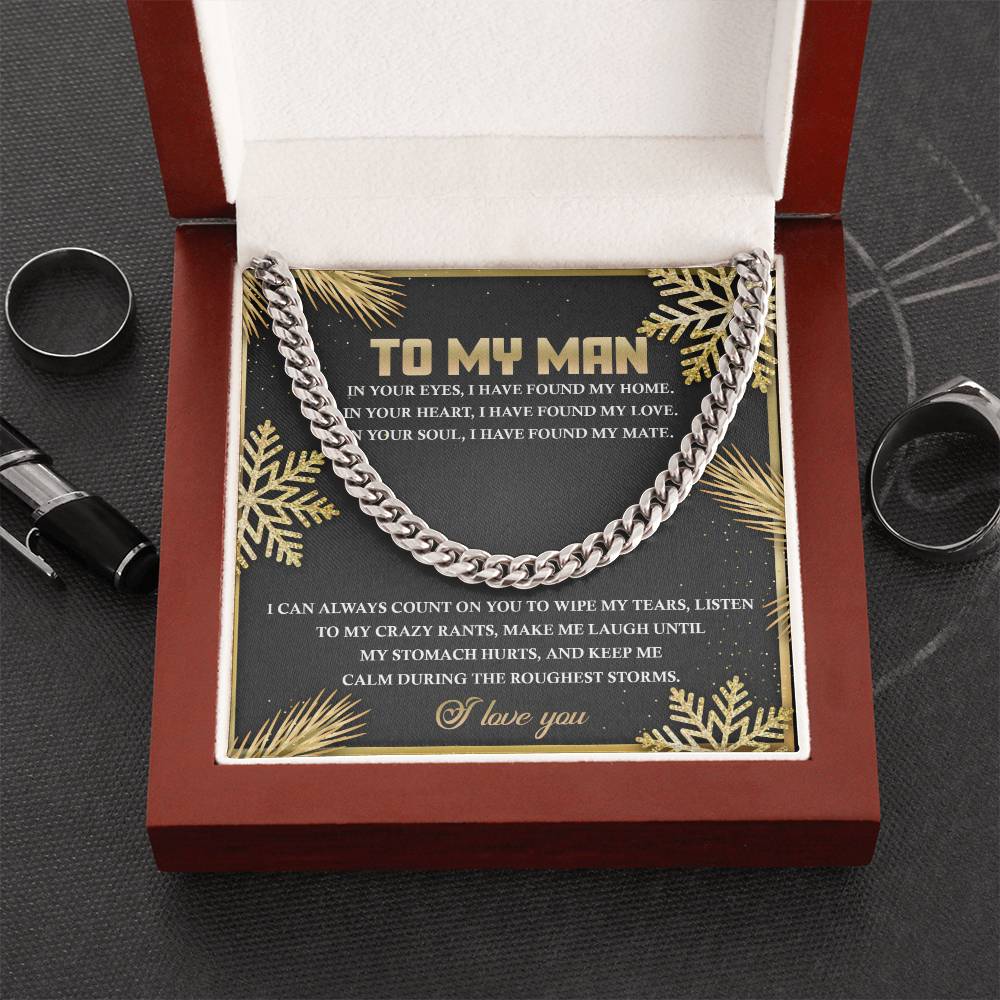 The "My Man-Found My Home - Cuban Link Chain" necklace, elegantly displayed in an open red box with a heartfelt message on the inner lid, rests gracefully. Surrounded by decorative elements and rings on a black surface, this personalized gift is ideal for any occasion.