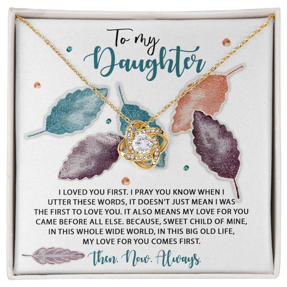 The Daughter-Pray You Know - Love Knot Necklace features a gold finish and a heart-shaped pendant adorned with sparkling cubic zirconia crystals. The necklace is placed on a beautifully designed card that reads: "To my Daughter. I loved you first..." surrounded by colorful leaf illustrations.